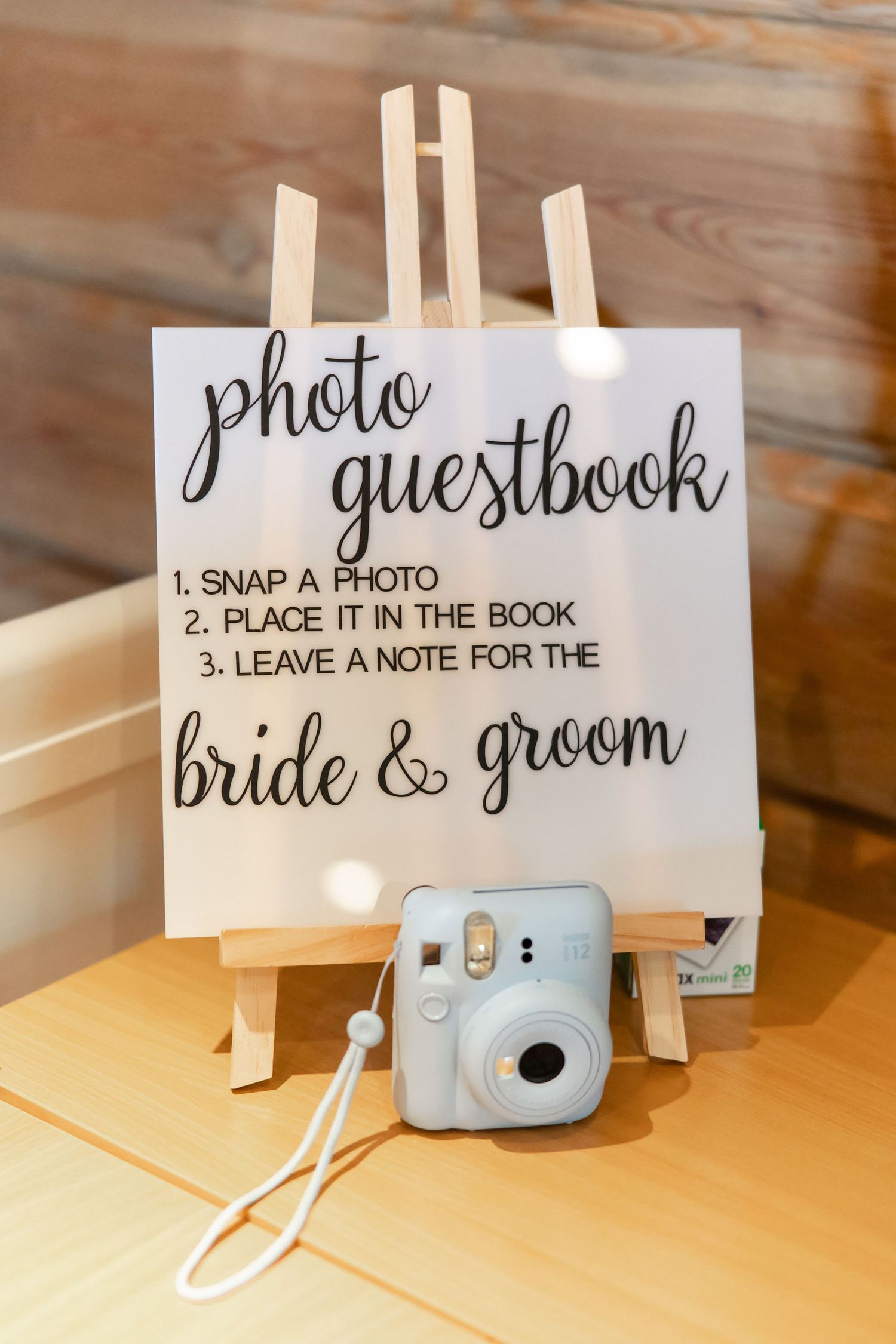 A series of wedding guestbook signs with a light blue Instax camera displayed on wooden easels against a rustic background.