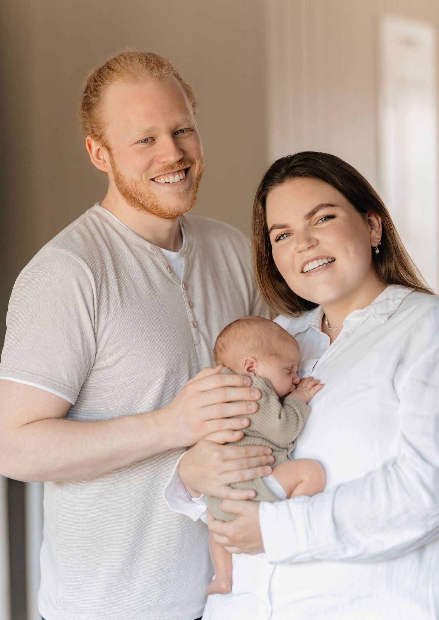 Smiling couple holding a sleeping newborn baby, posing together for a family portrait in a bright room.