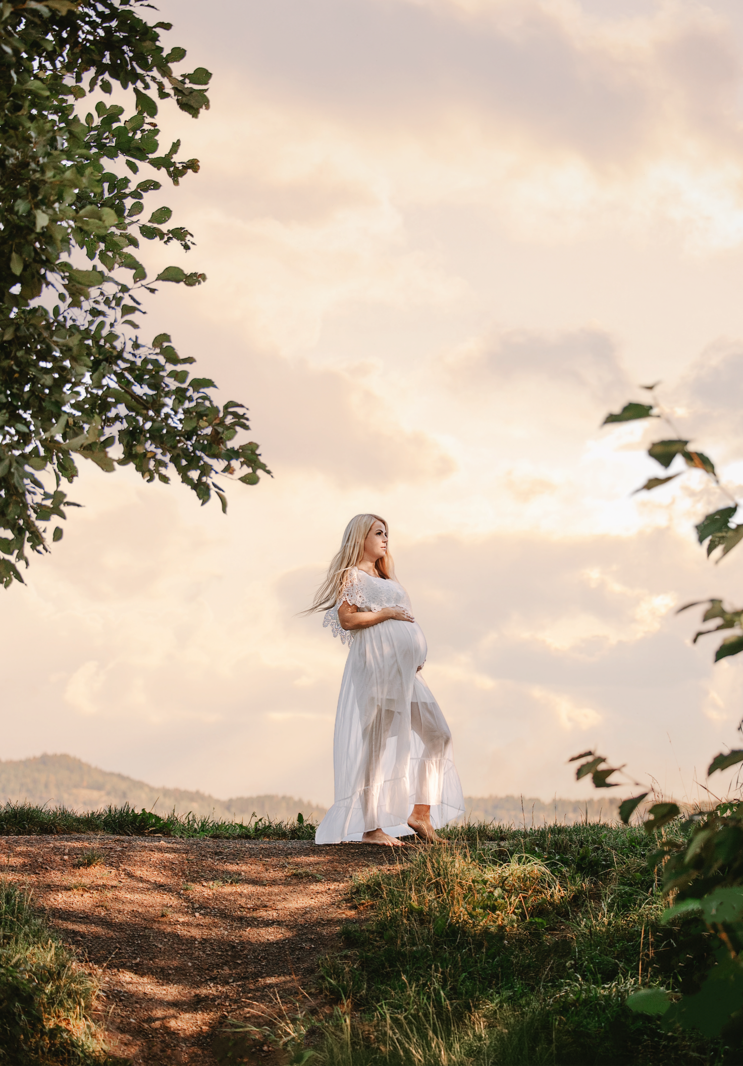 A person in a flowing white dress stands against a dramatic sunset sky on a hilltop with green foliage.