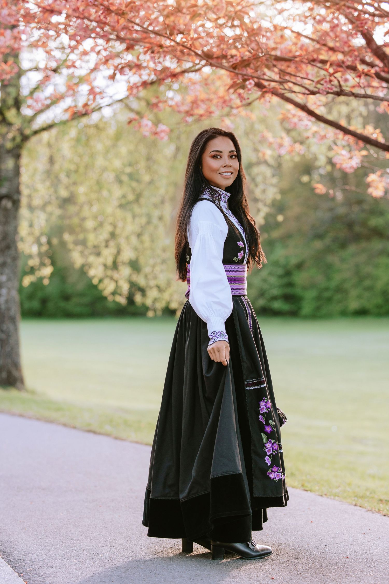 A woman in a black velvet dress and white blouse poses under blooming pink cherry blossoms in spring.