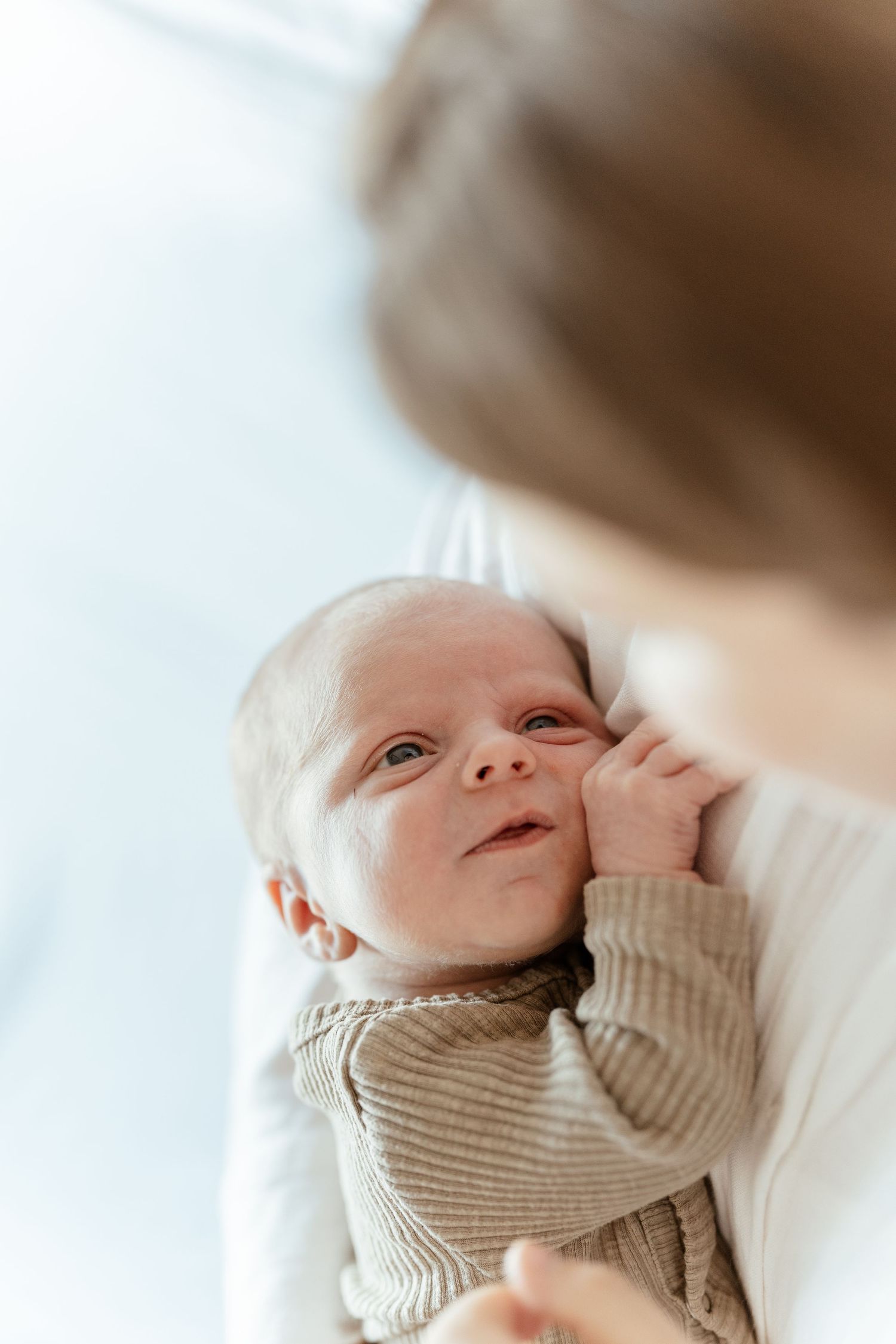 Newborn baby in beige outfit gazing up at mother, cradled in her arms against a bright white background.