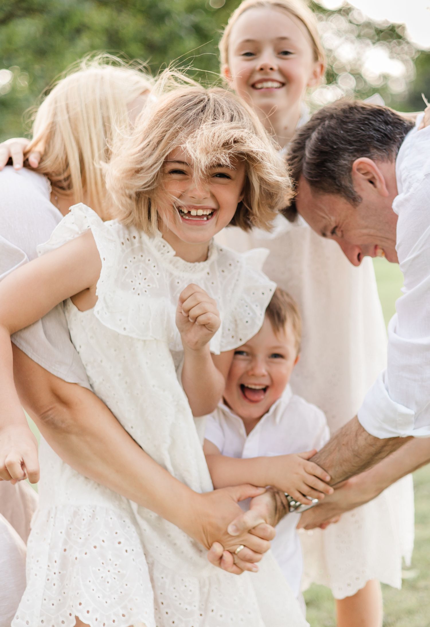 A joyful family wearing white outfits shares candid laughing moments together during an outdoor photo session.