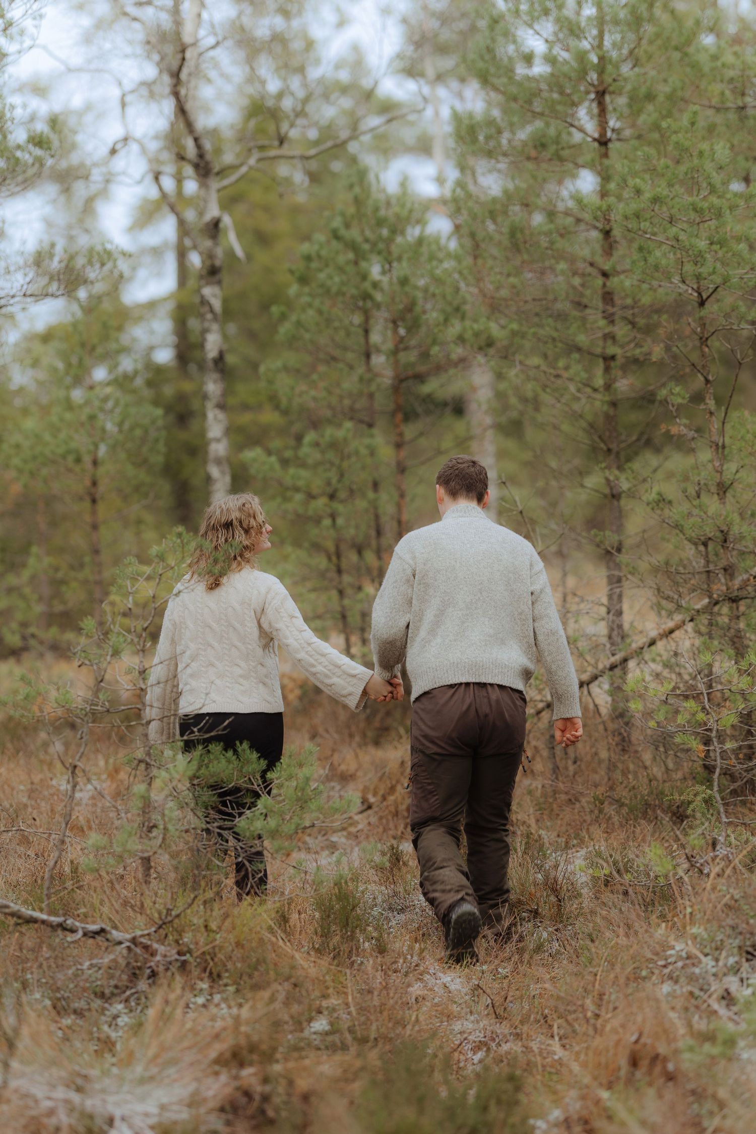 Two people walk through a winter forest with bare trees and dried brush on the ground.