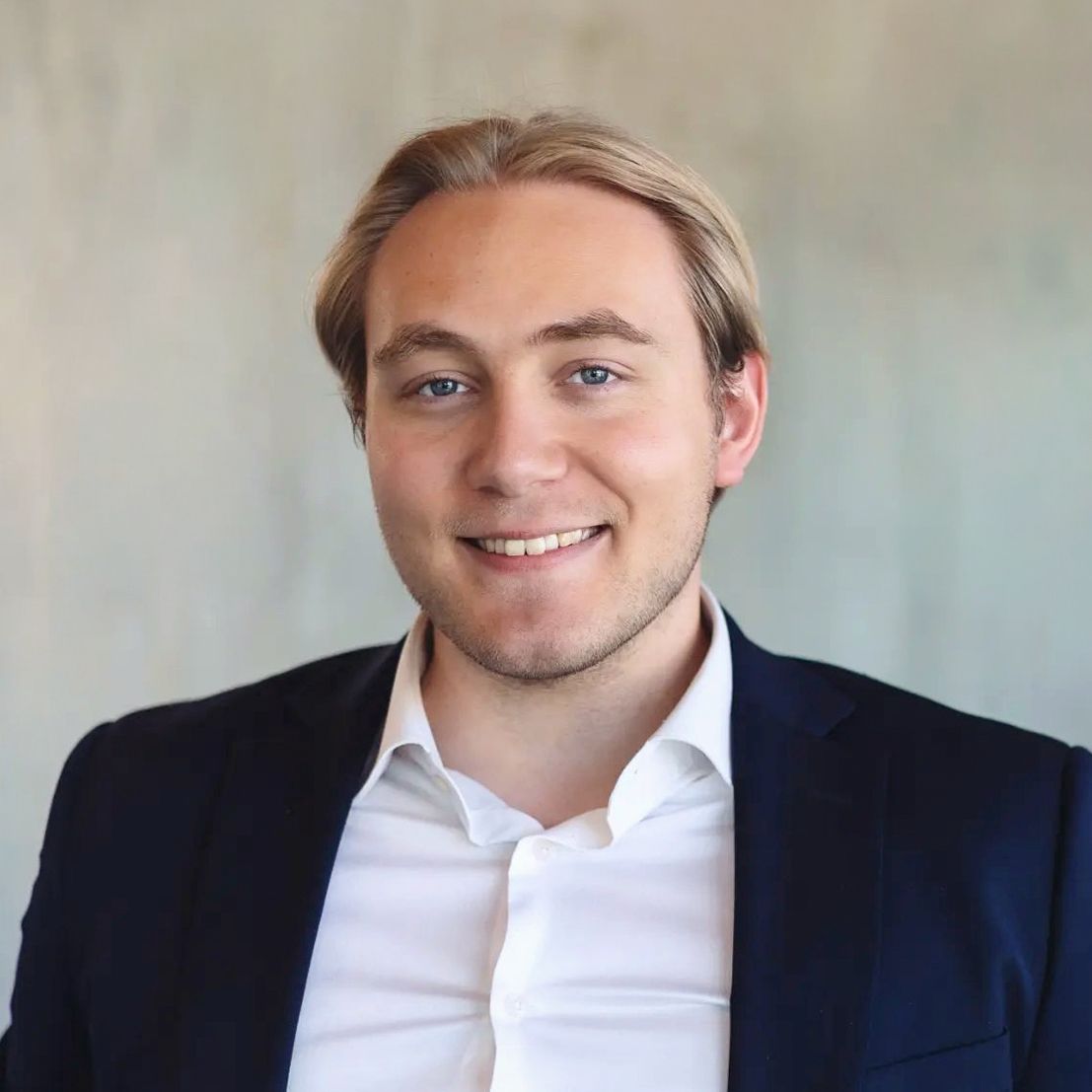 Professional headshot of someone wearing a navy suit jacket and white dress shirt against a light background.