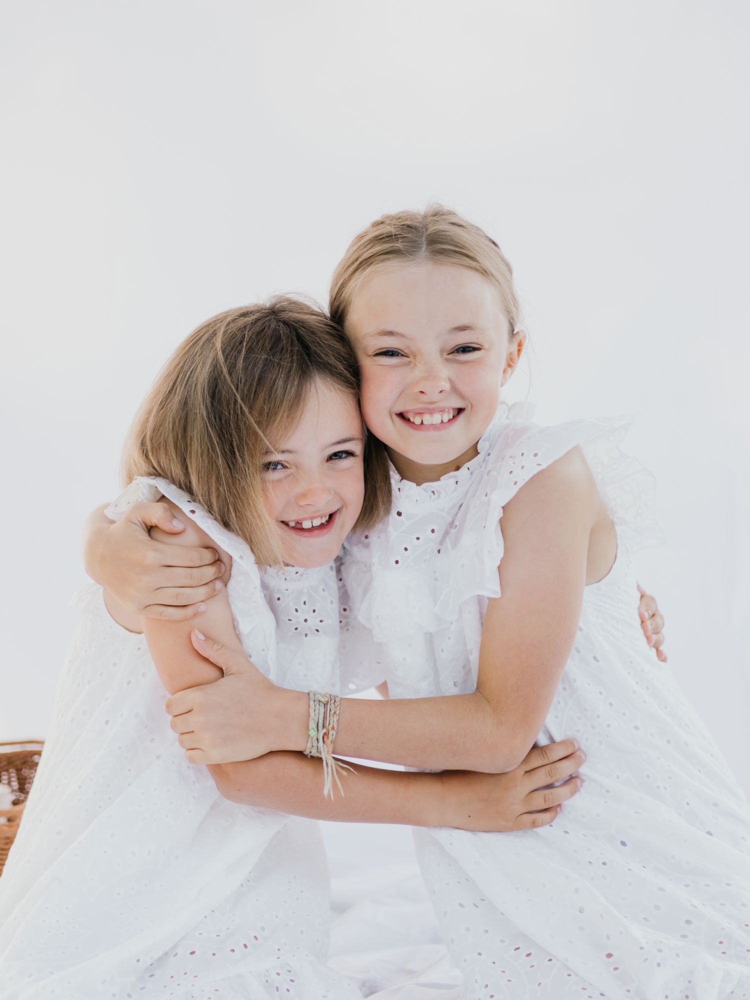 Two children in white dresses share joyful hugs and laughter in a bright studio setting.