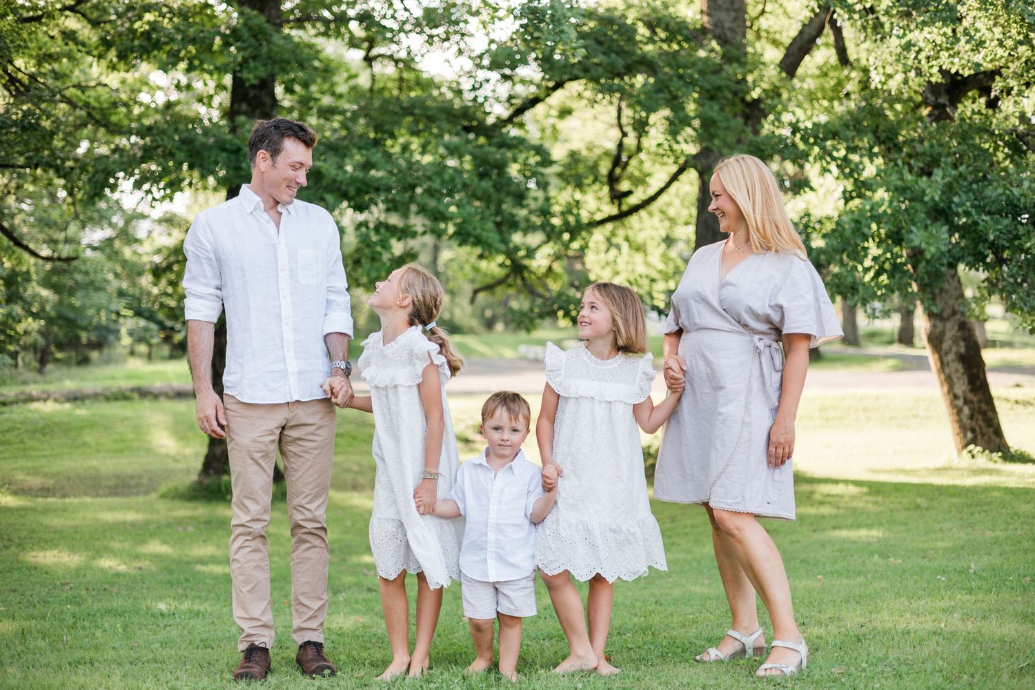 A group wearing white and beige outfits walks together through a sunlit park on a summer day.