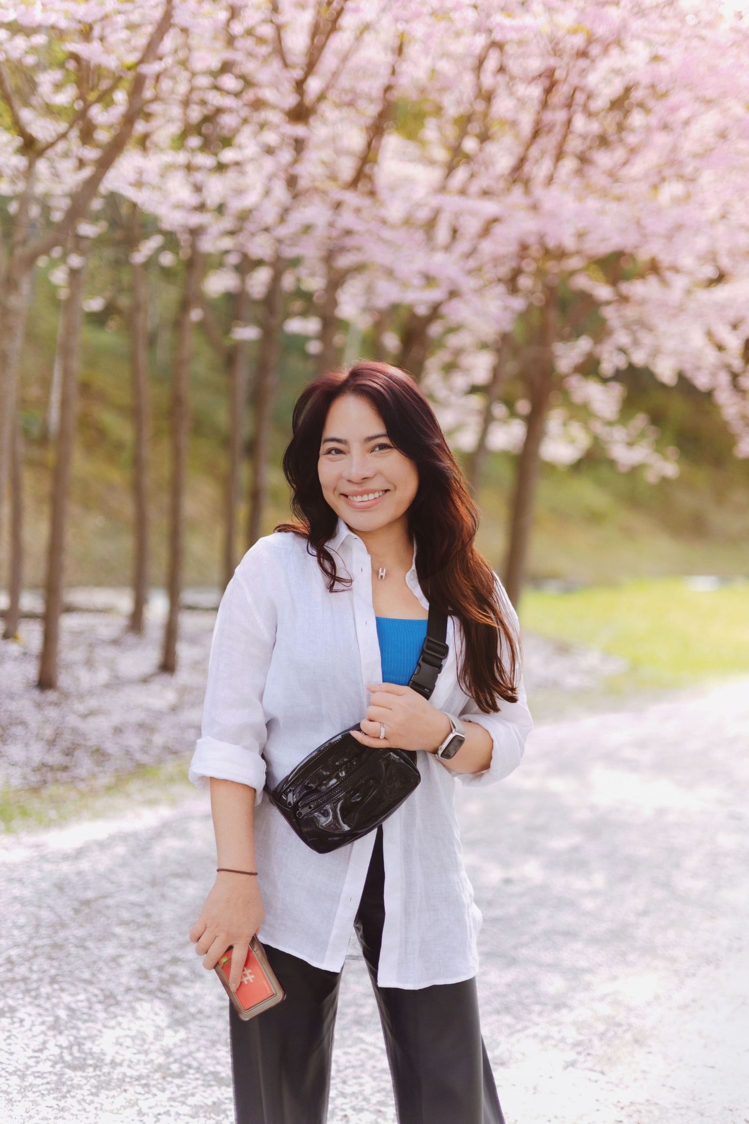 Woman in white jacket poses under blooming cherry blossom trees in spring park setting.