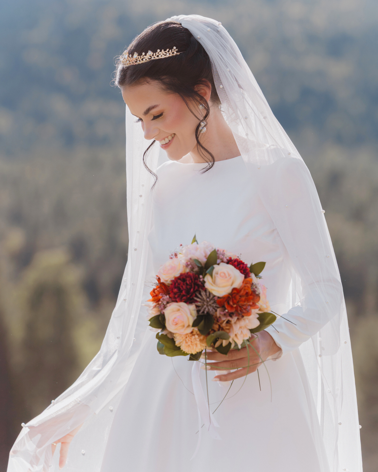 A bride in a classic white long-sleeved wedding dress holds a vibrant autumn bouquet against a mountain backdrop.
