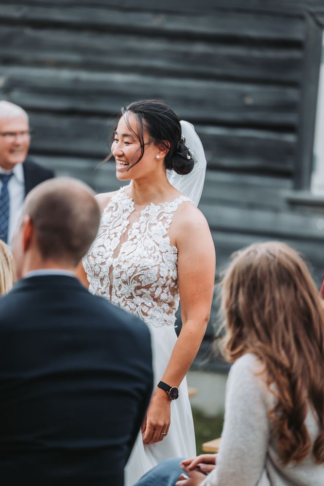A bride in a white lace wedding dress laughs joyfully during an outdoor ceremony.