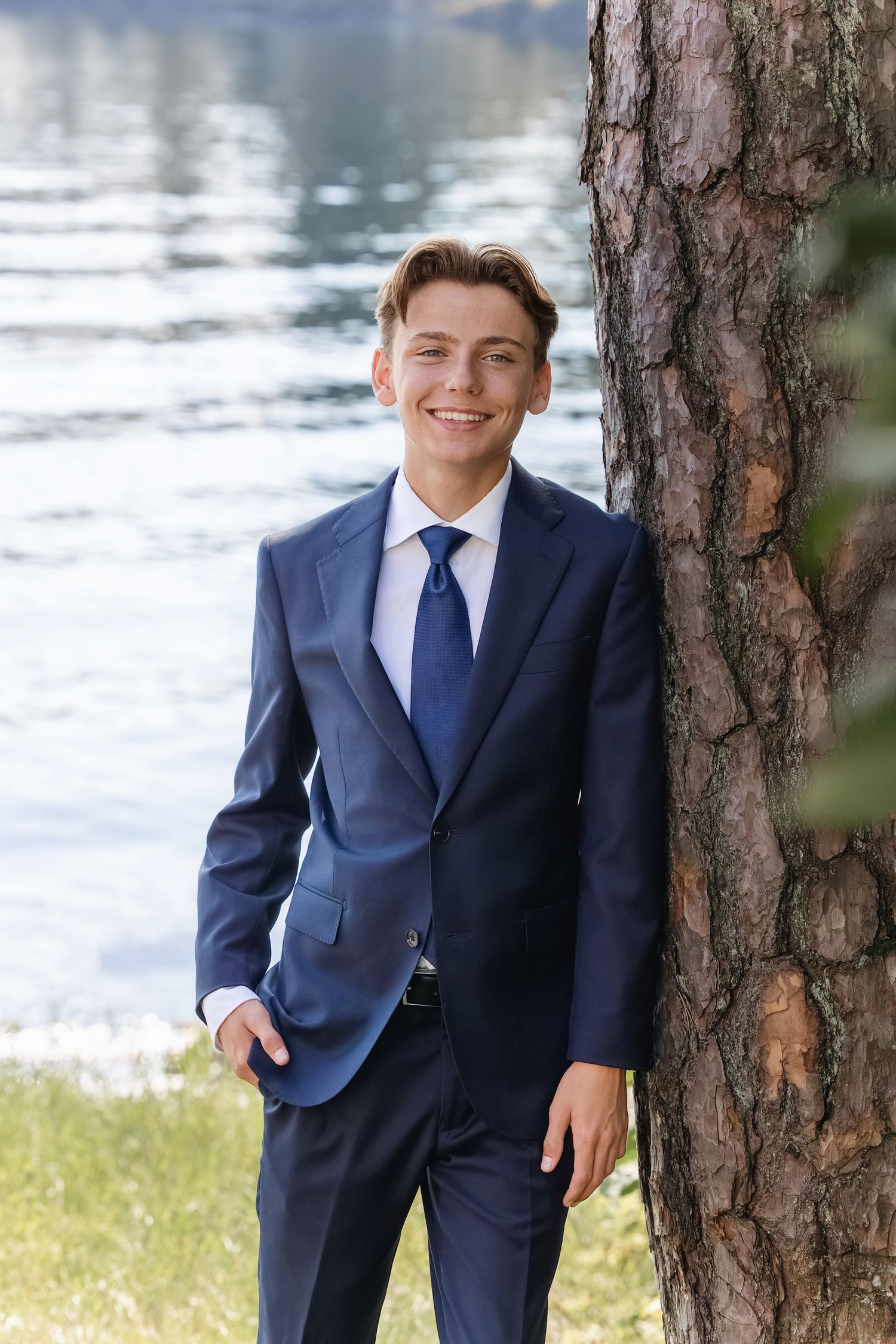 Young person in a navy blue suit poses casually next to a tree trunk by a lake in an outdoor setting.
