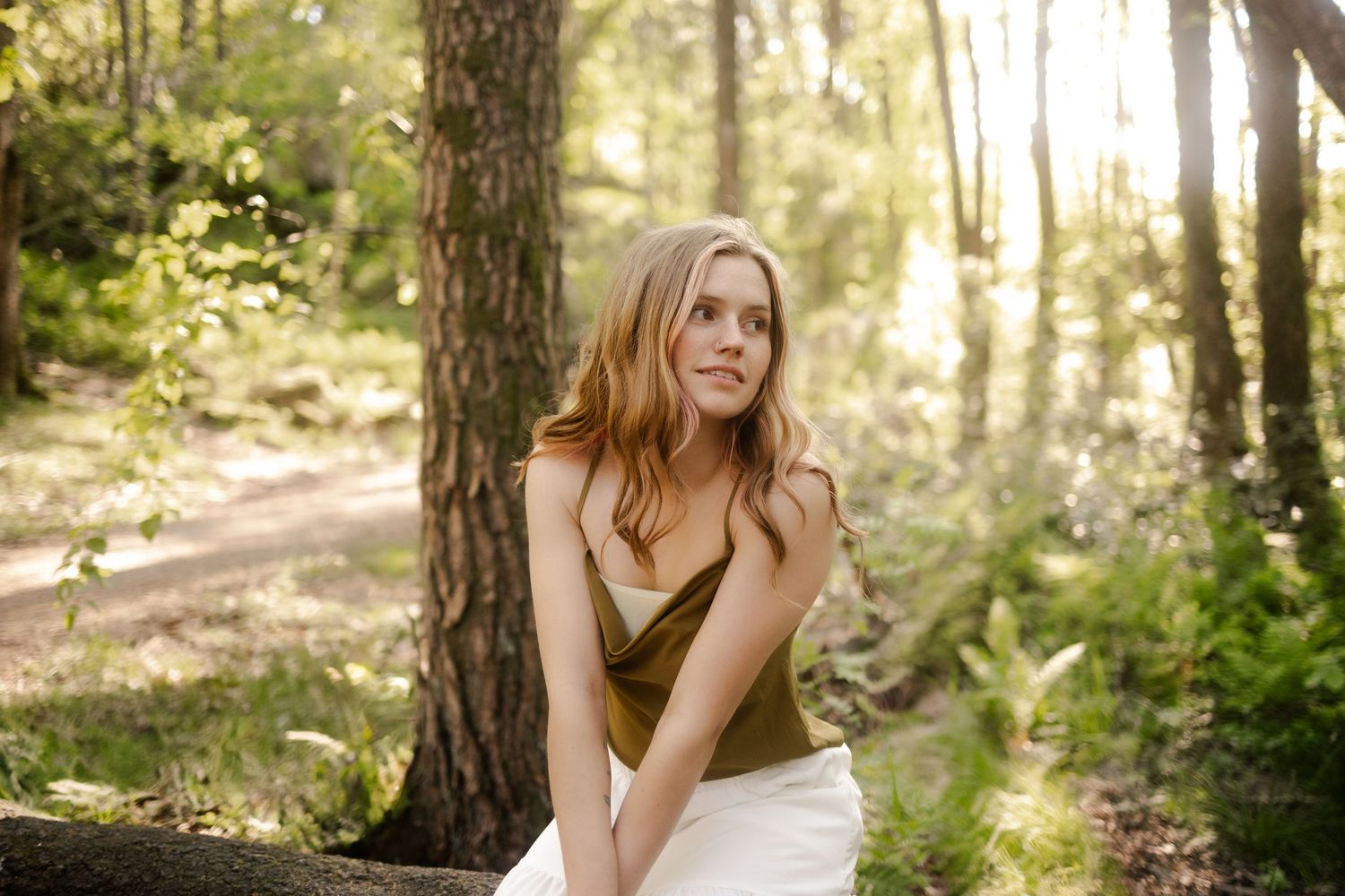 A person poses in a sunlit forest wearing a white dress during an outdoor portrait photography session.