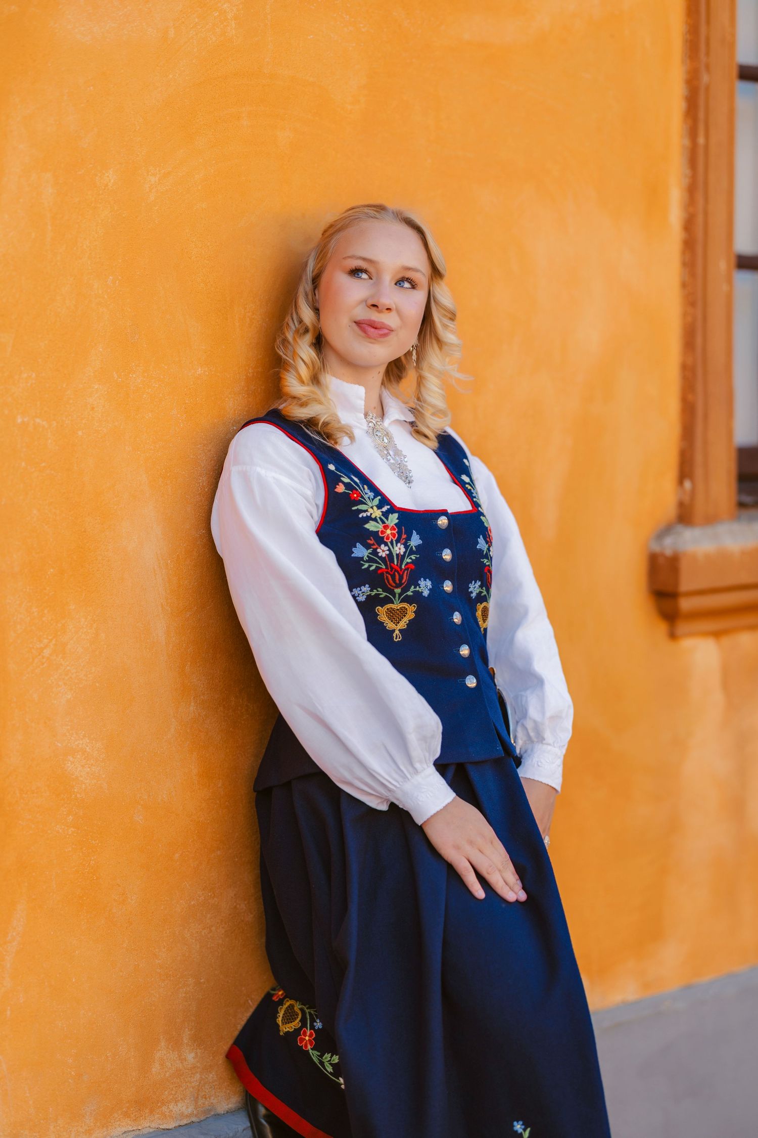 Young blonde woman in traditional Norwegian bunad folk costume posing against a vibrant orange wall.