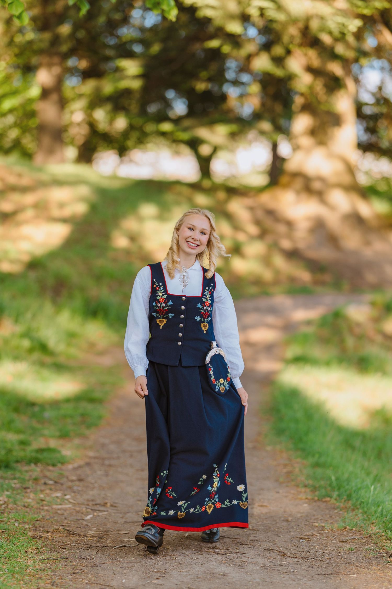Young blonde woman in traditional Norwegian bunad folk costume walking on a dirt path surrounded by green trees.