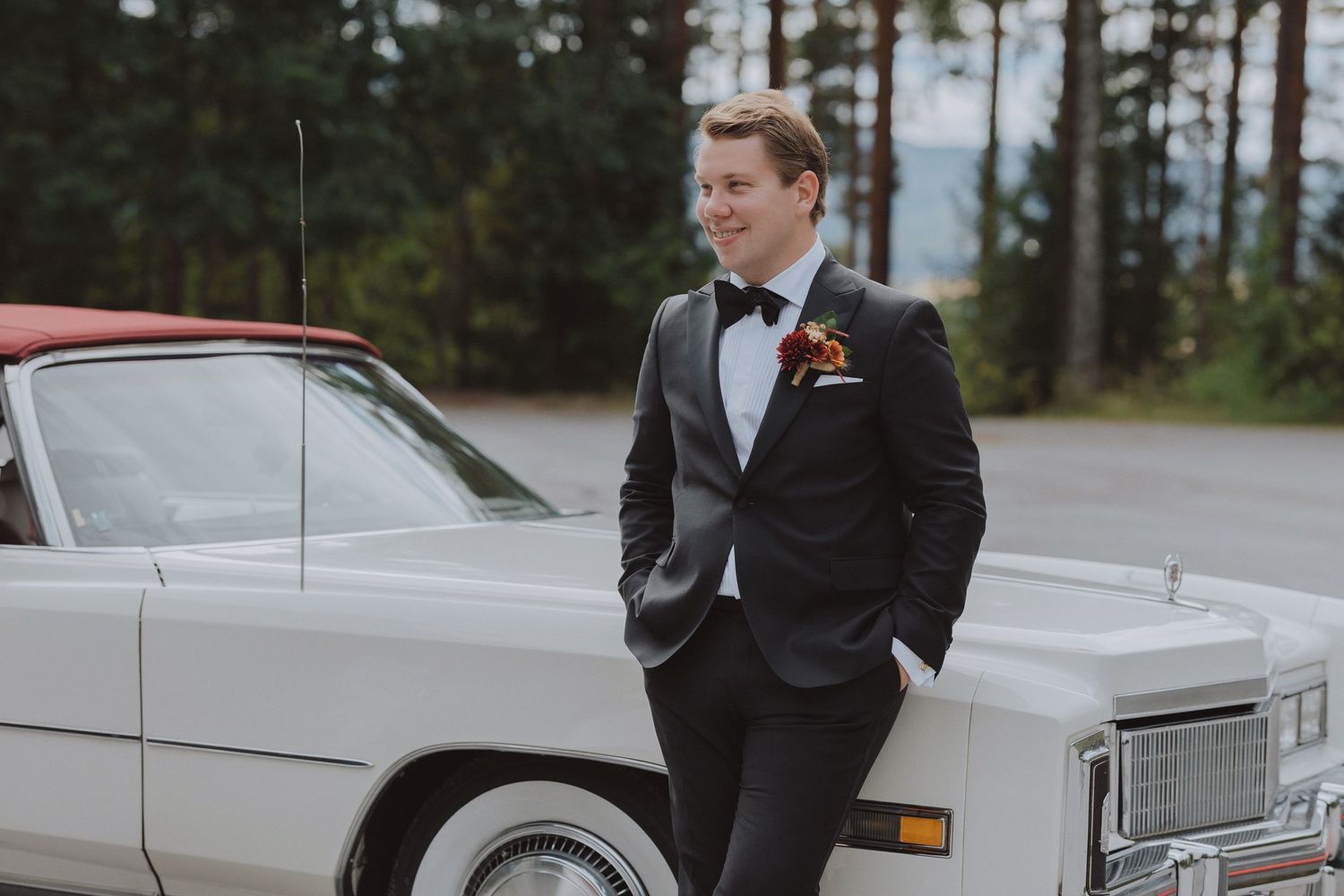 A wedding party poses with classic white vintage cars in a forest setting.