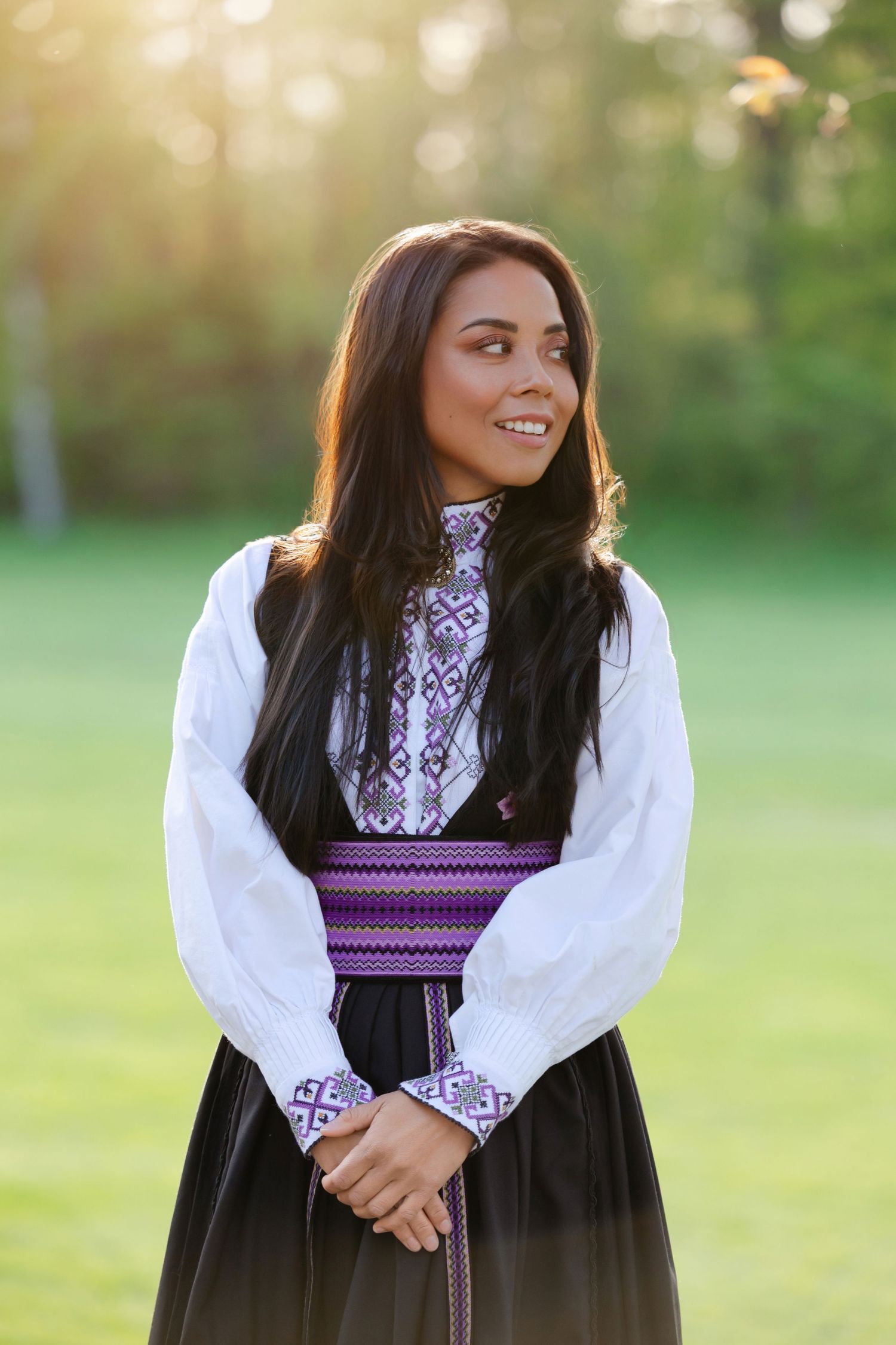 A woman in traditional dress with white sleeves and purple embroidered details poses outdoors in a grassy field.