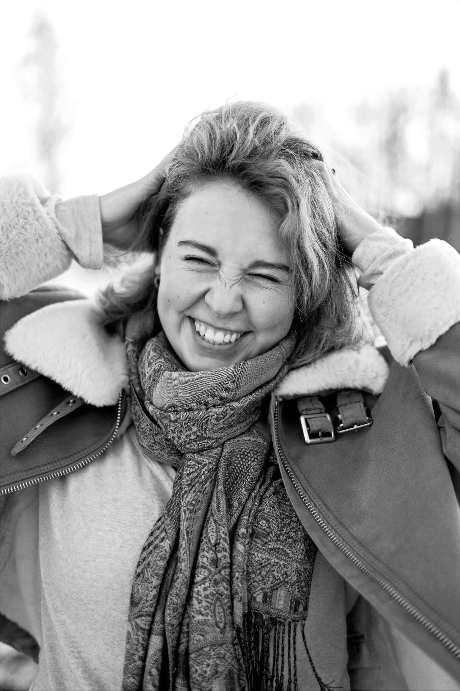 Black and white portrait of someone in a winter coat and scarf, hands in hair, laughing joyfully outdoors.