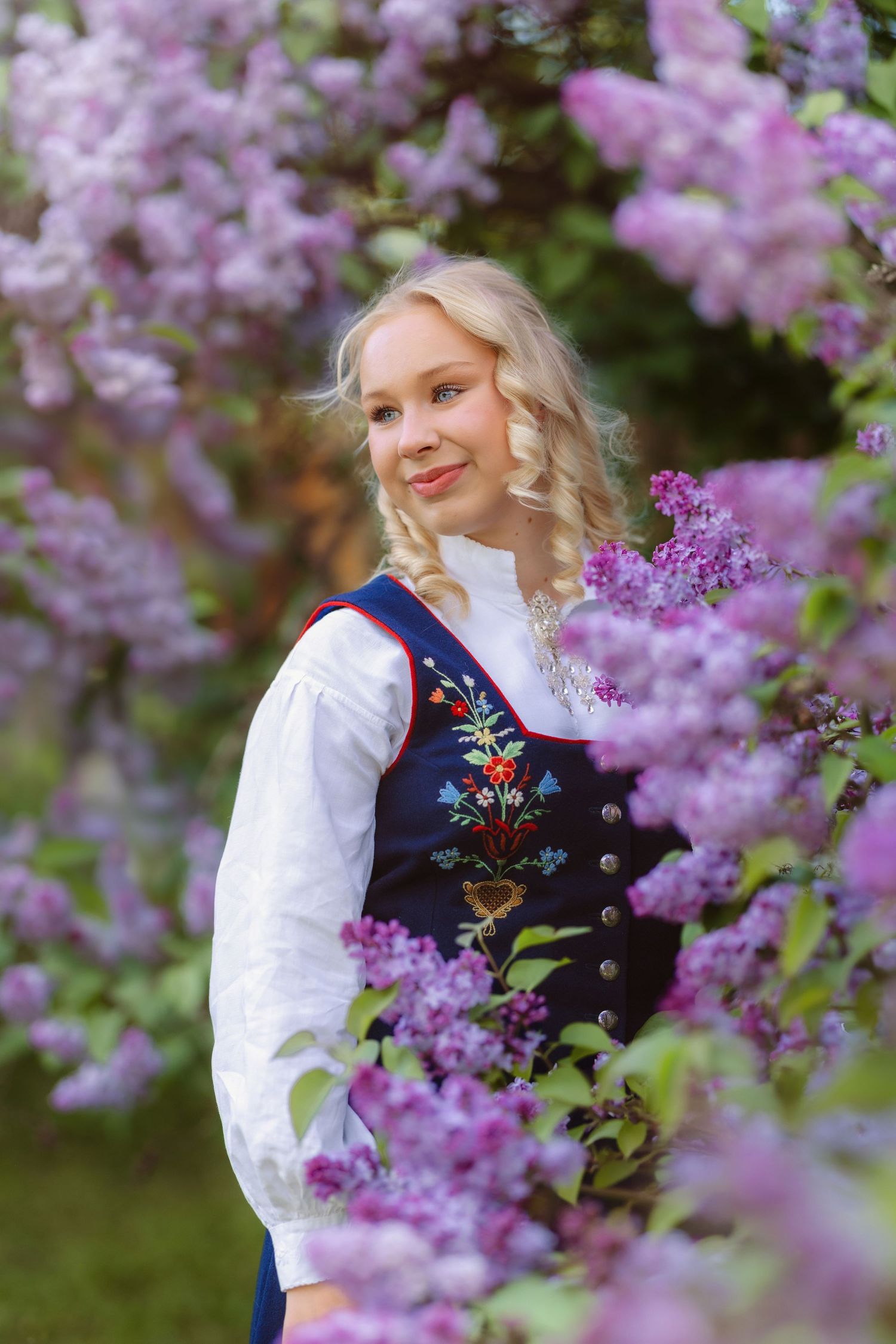 Blonde girl in Norwegian bunad traditional dress surrounded by blooming purple lilac flowers in a garden.