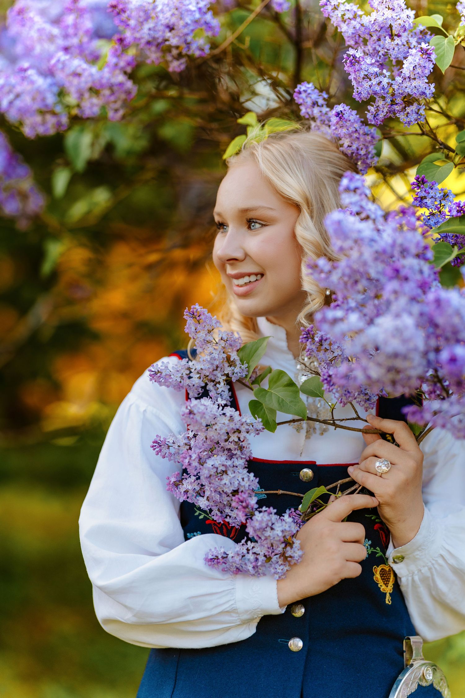 Smiling blonde woman in traditional Nordic dress holding purple lilac flowers outdoors in golden light.
