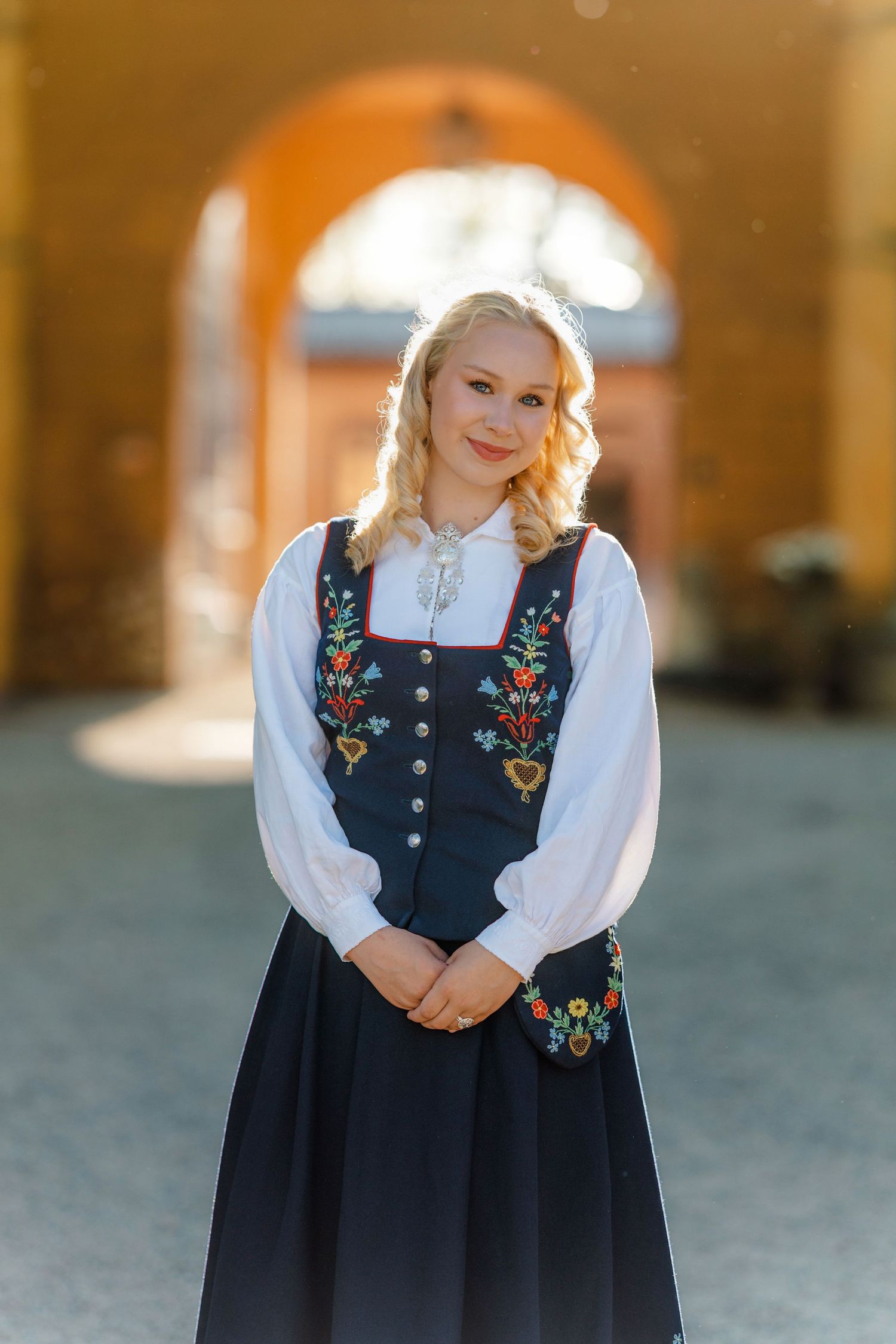 Young blonde woman in traditional Norwegian bunad dress with floral embroidery, smiling outdoors in golden light.