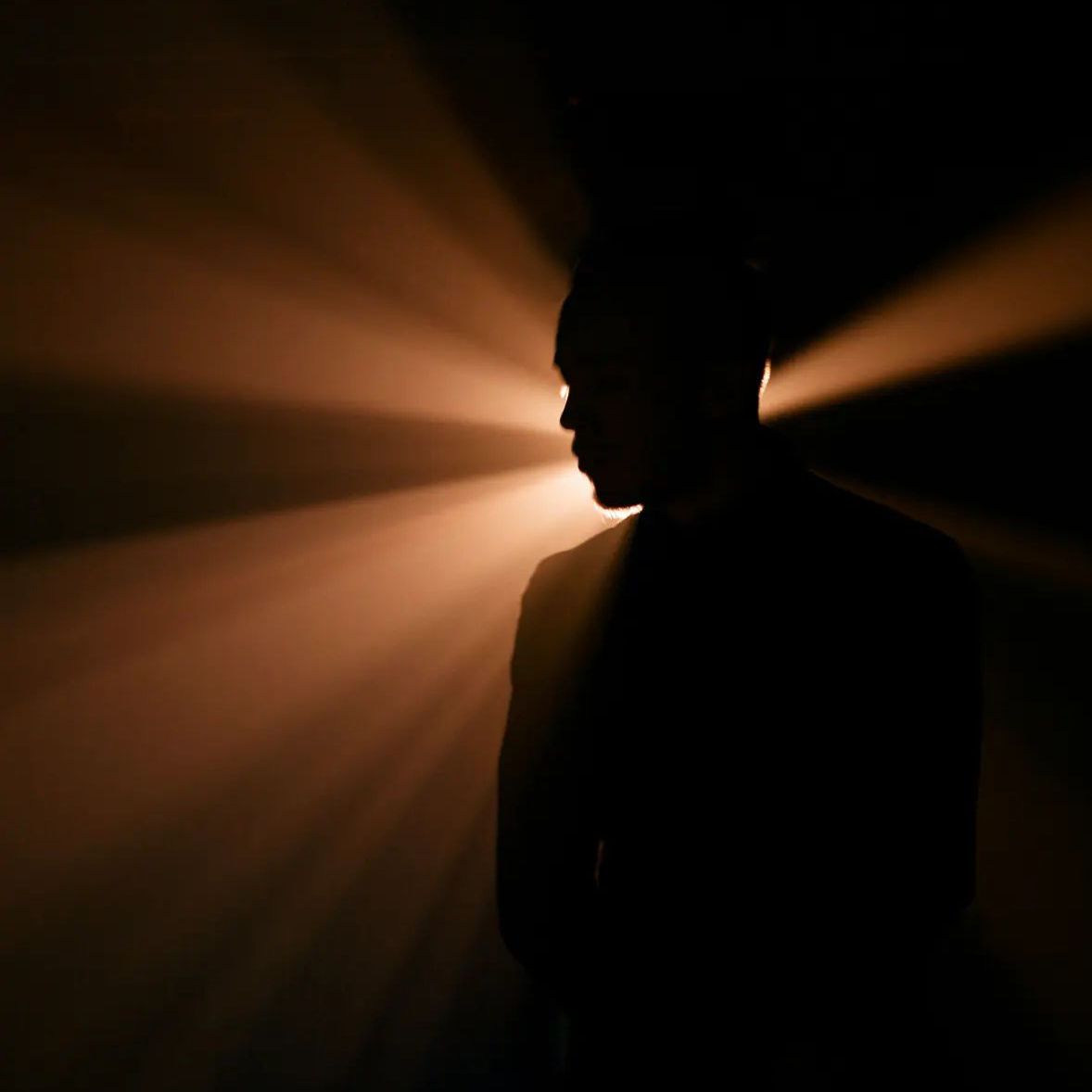 Silhouette of a person's head against a dark background with warm orange light rays emanating from behind.
