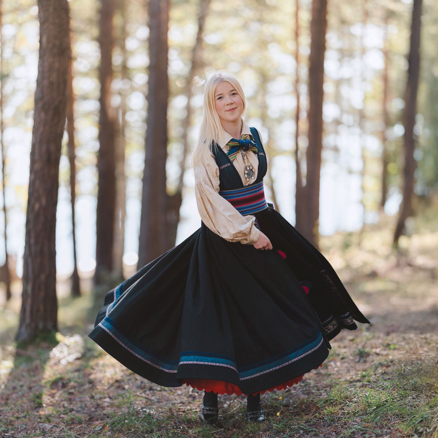 Traditional Norwegian folk costume dress with flowing black skirt twirls in a sunlit forest setting.