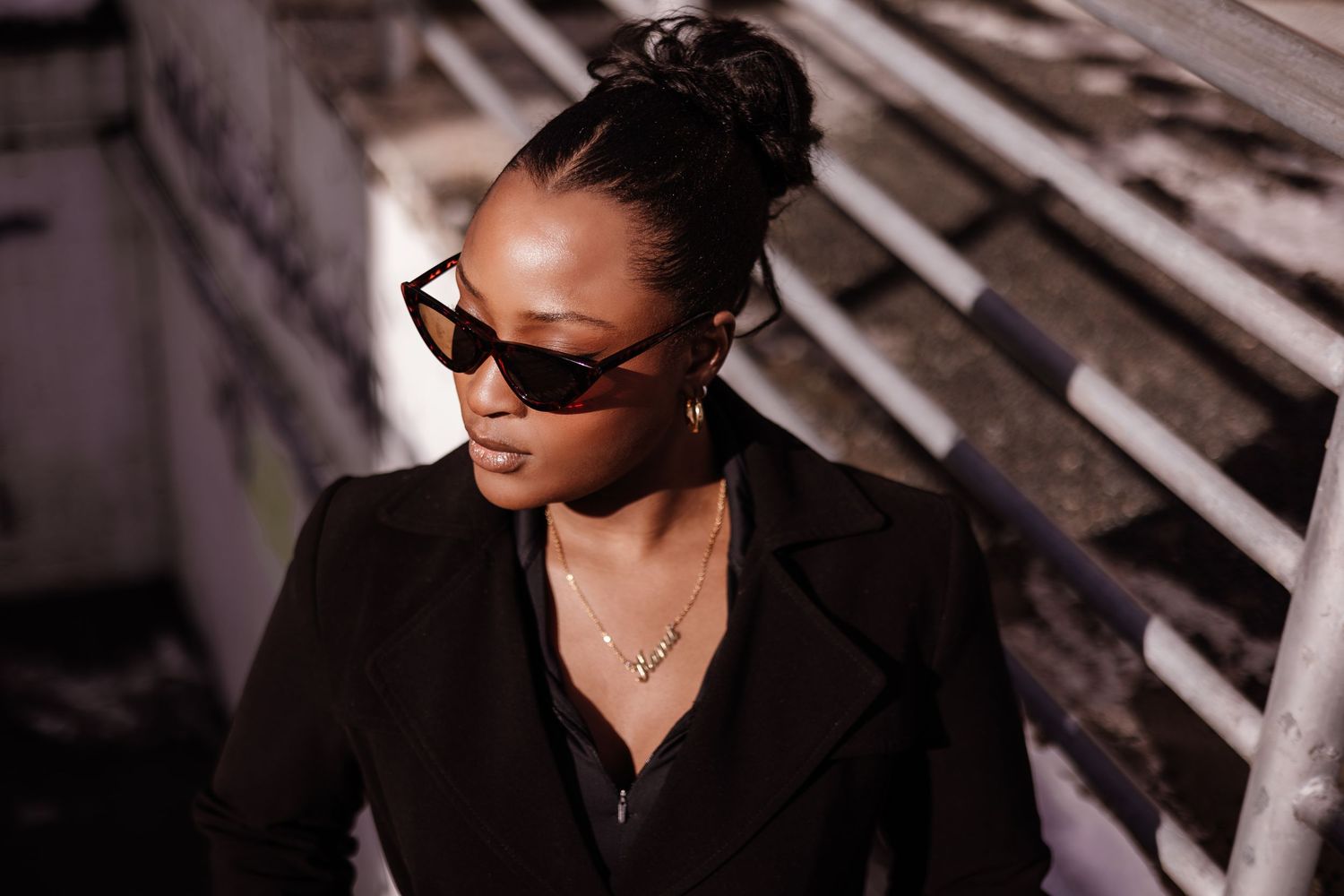 Person wearing sunglasses and dark clothing poses against wooden slat background in dramatic lighting.