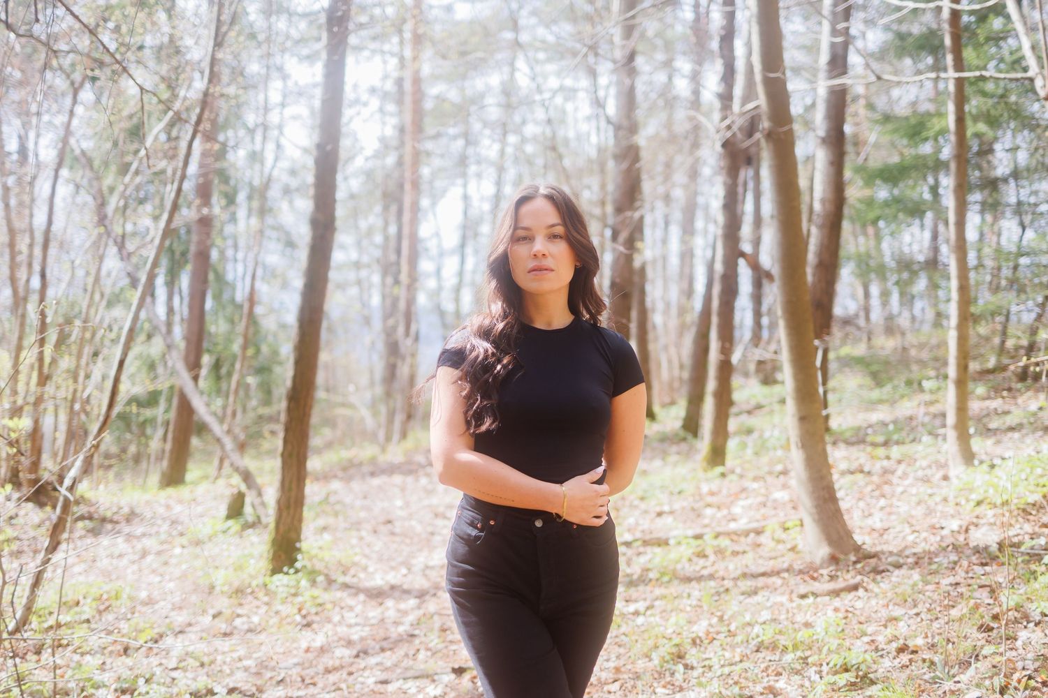 A person in a black top stands in a wooded forest area surrounded by tall trees and fallen leaves.