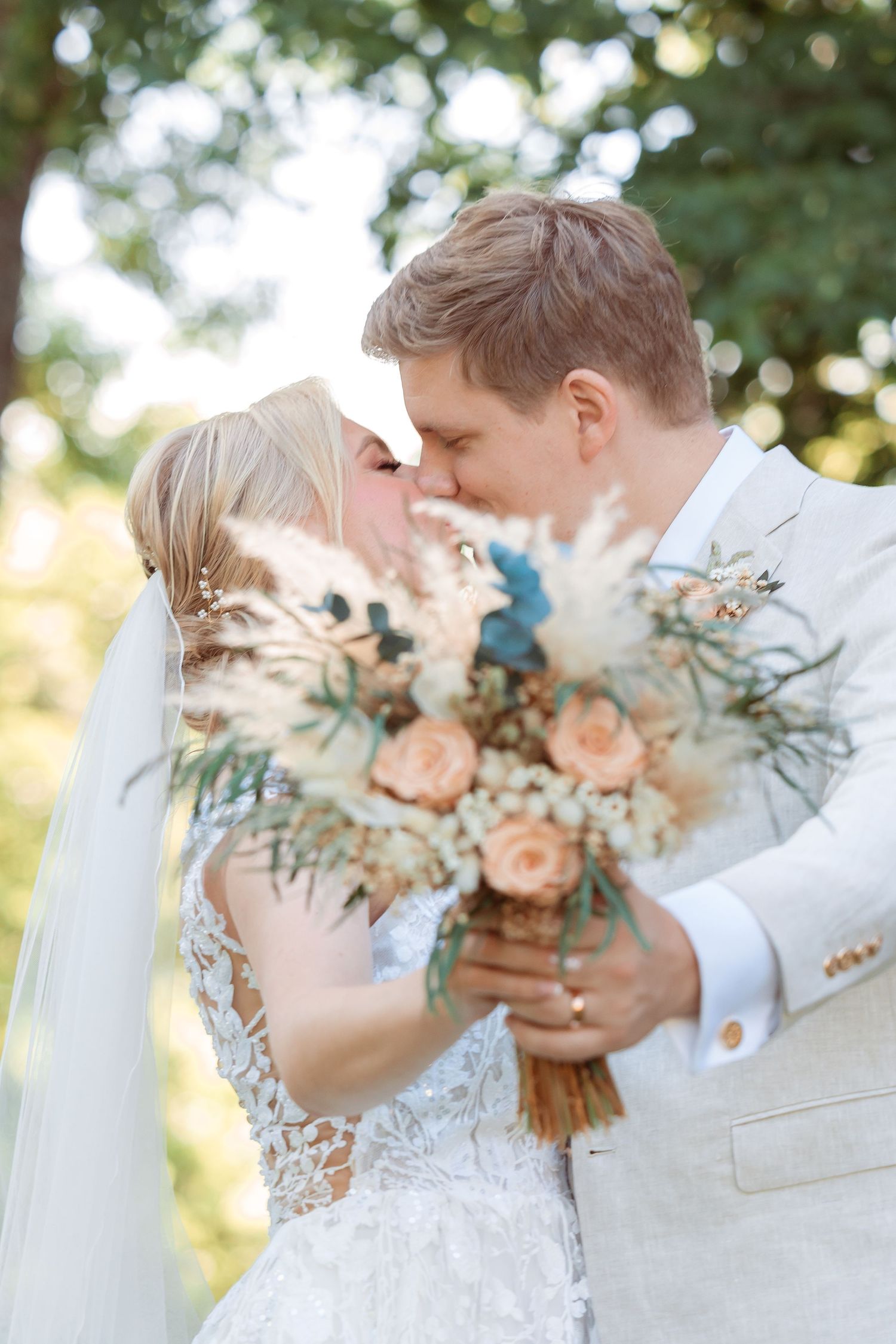 A romantic wedding couple shares tender moments behind a peach and cream floral bouquet in an outdoor setting.