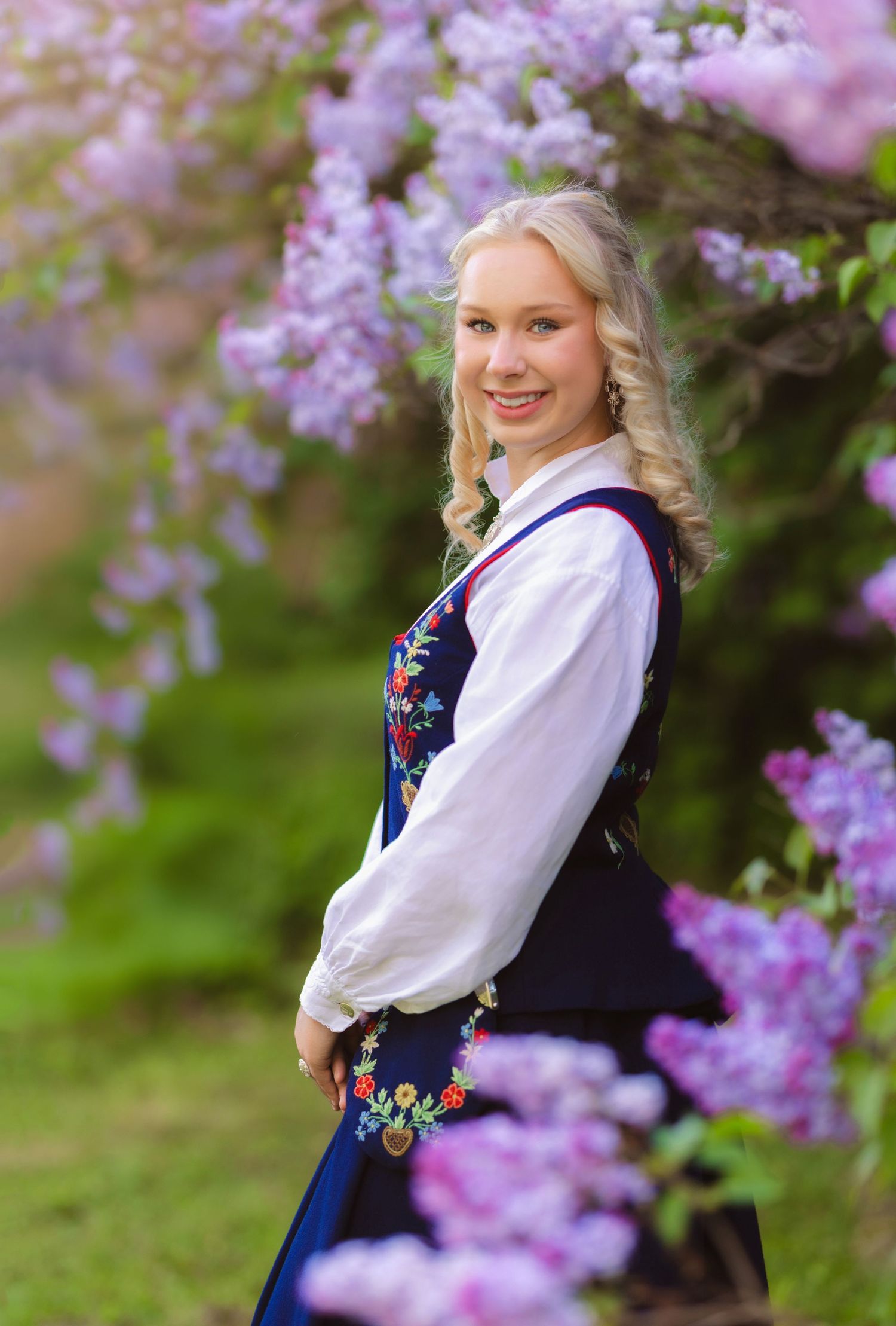 Young blonde woman in traditional Norwegian bunad dress smiles among blooming purple lilac flowers outdoors.