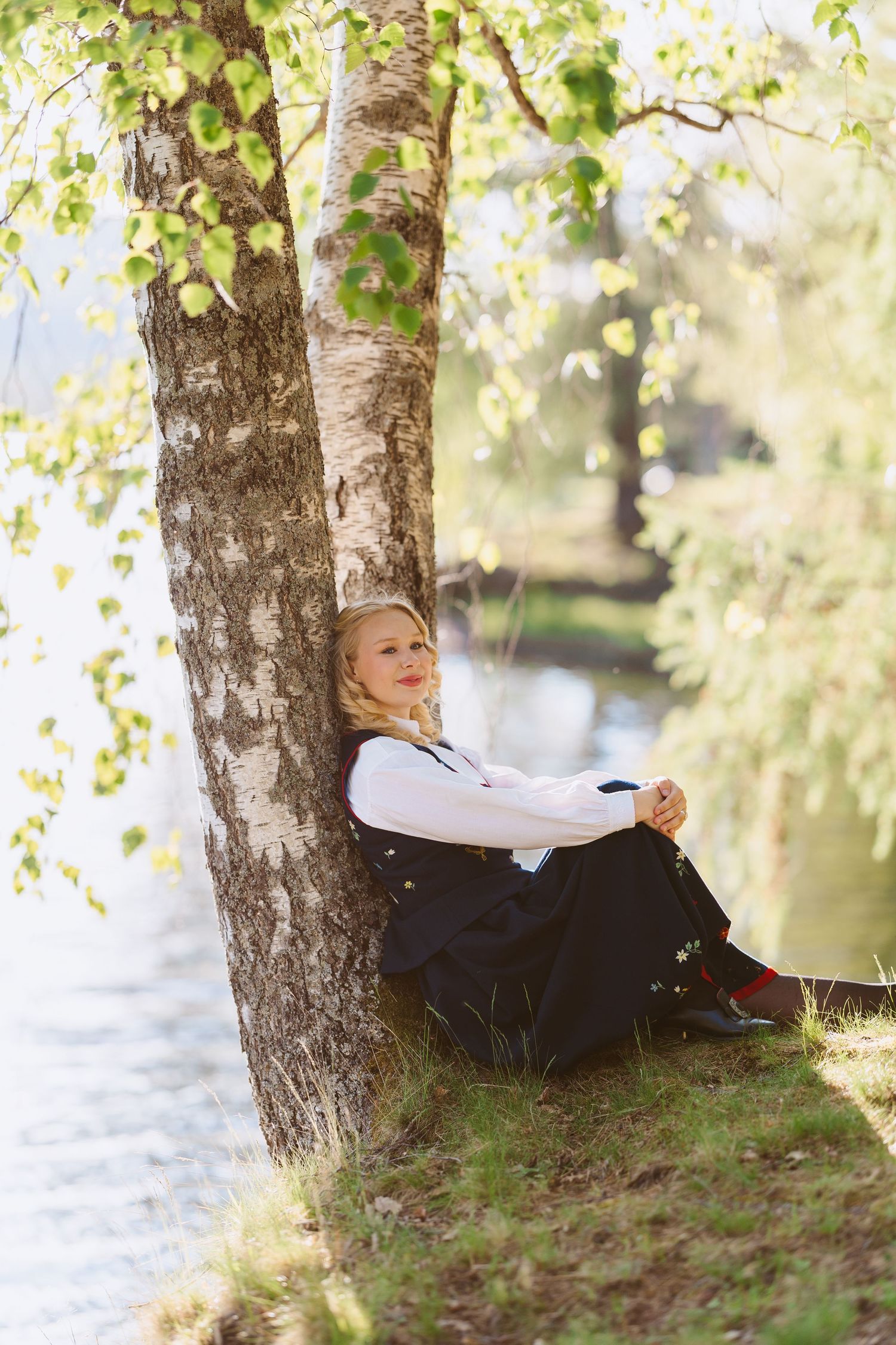 Blonde woman in white blouse and black skirt sitting against birch tree by a serene lake on a sunny day.