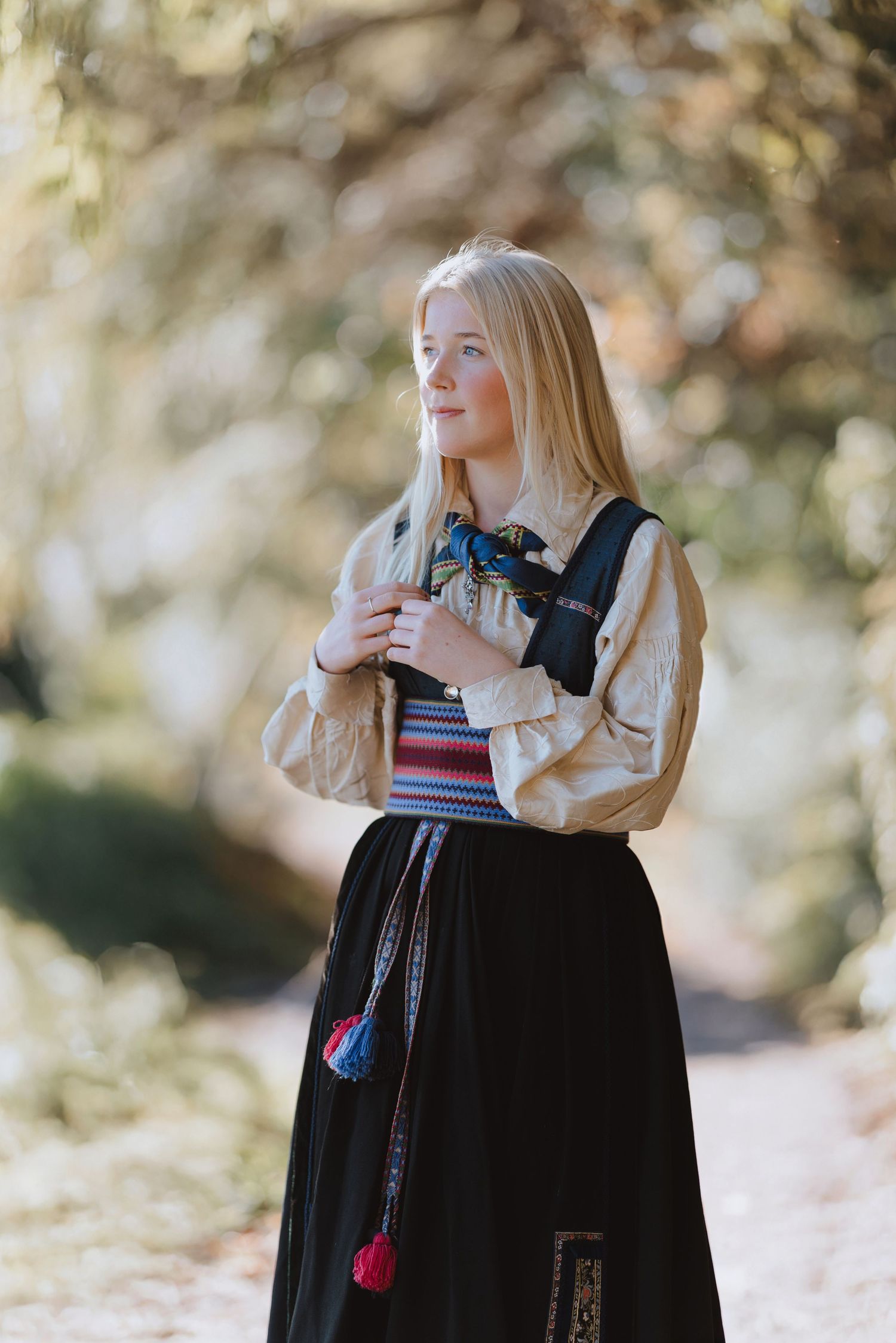 A person in a black cloak and light-colored top poses thoughtfully in an autumn outdoor setting with bokeh background.