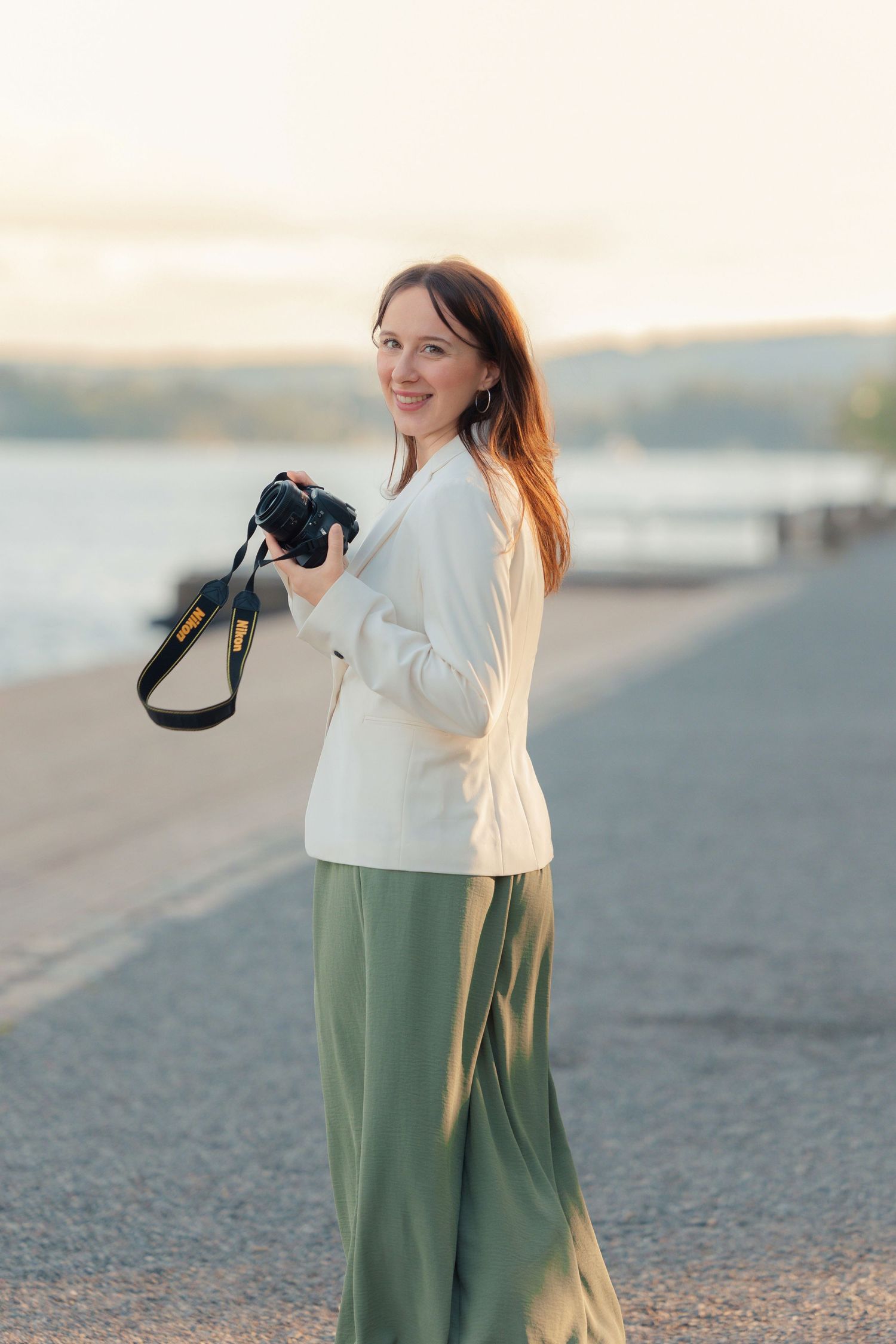 Photographer in white blazer and green skirt holding camera on waterfront path.
