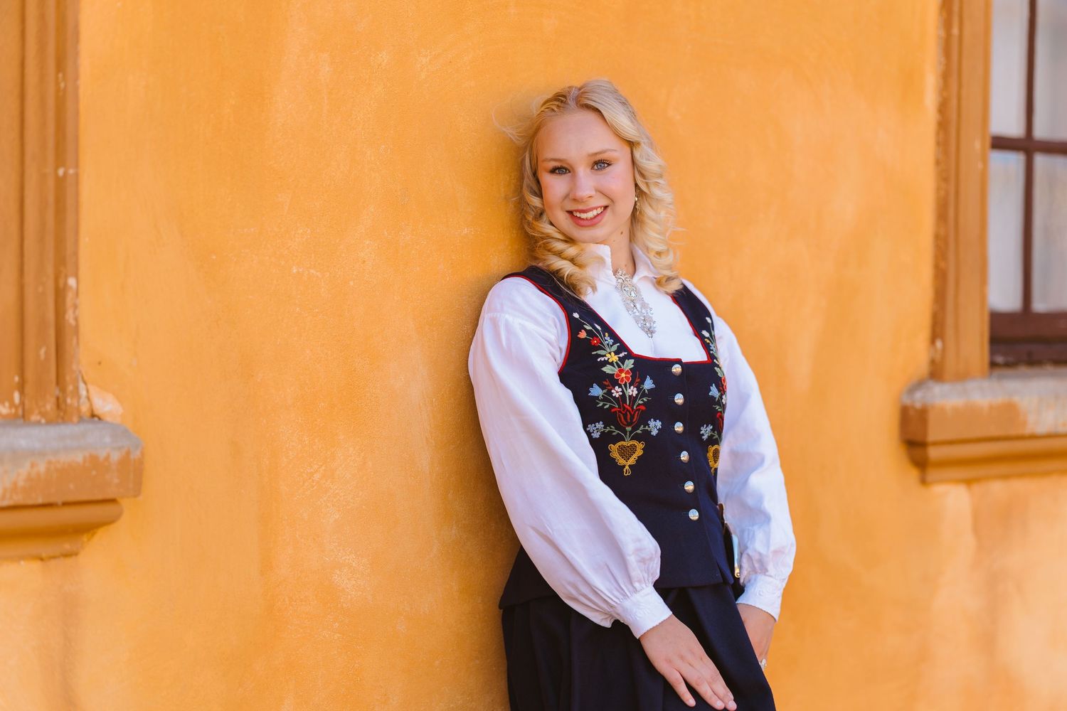 Young blonde woman in traditional Nordic folk costume with embroidered vest smiling against orange wall.