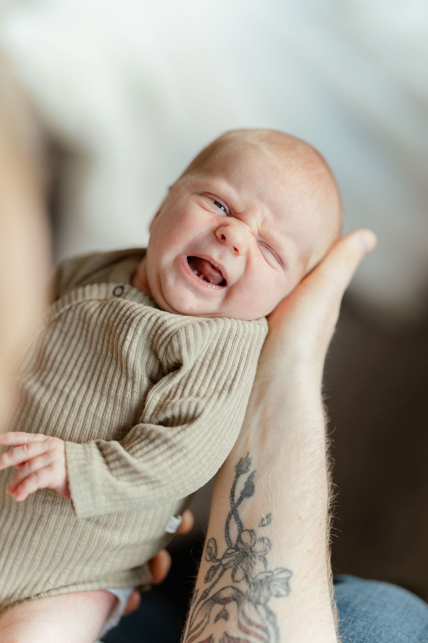 A crying newborn baby in a sage green ribbed onesie cradled in tattooed hands.