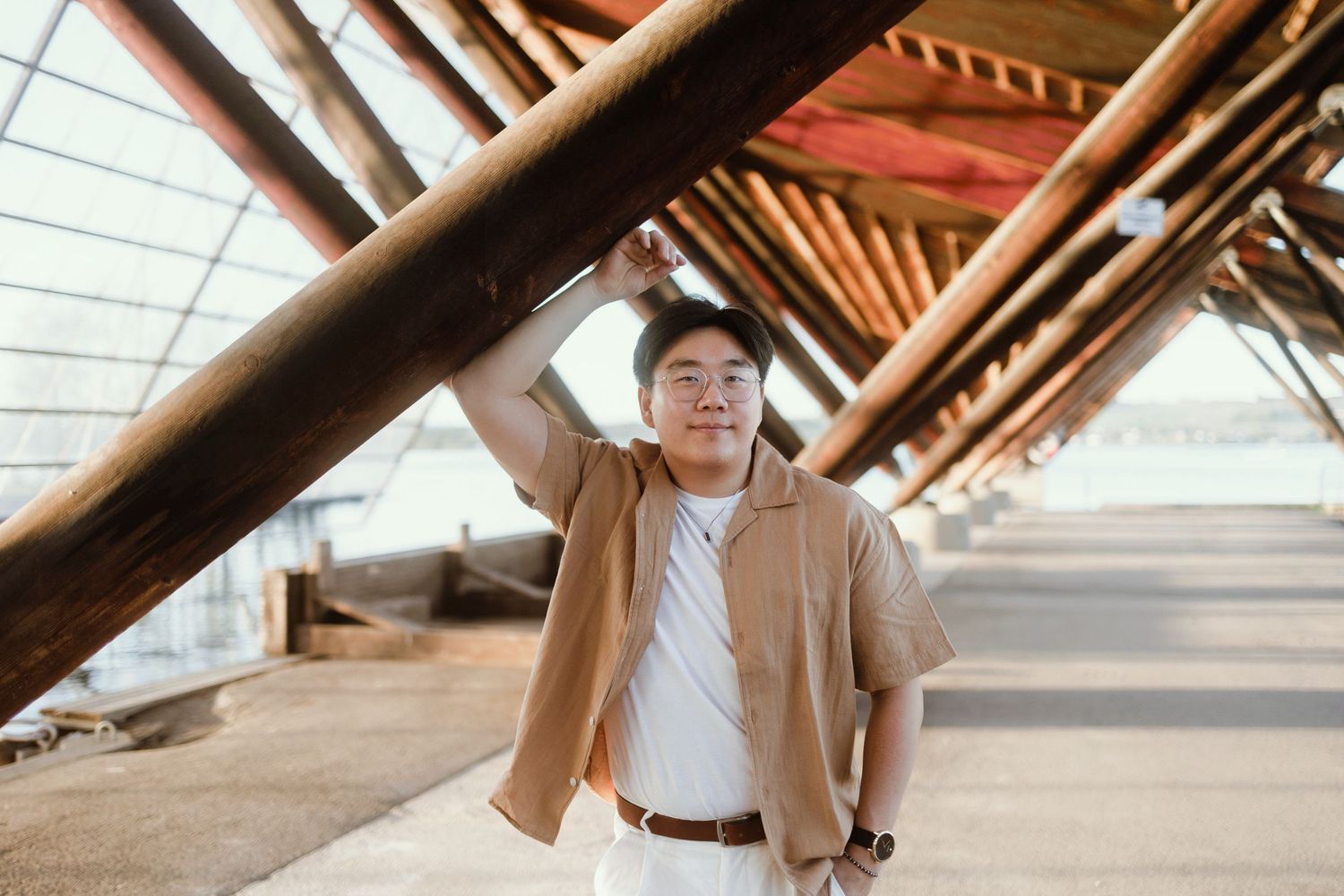 Person in tan jacket and white hat stands beneath angular wooden beams in architectural structure.