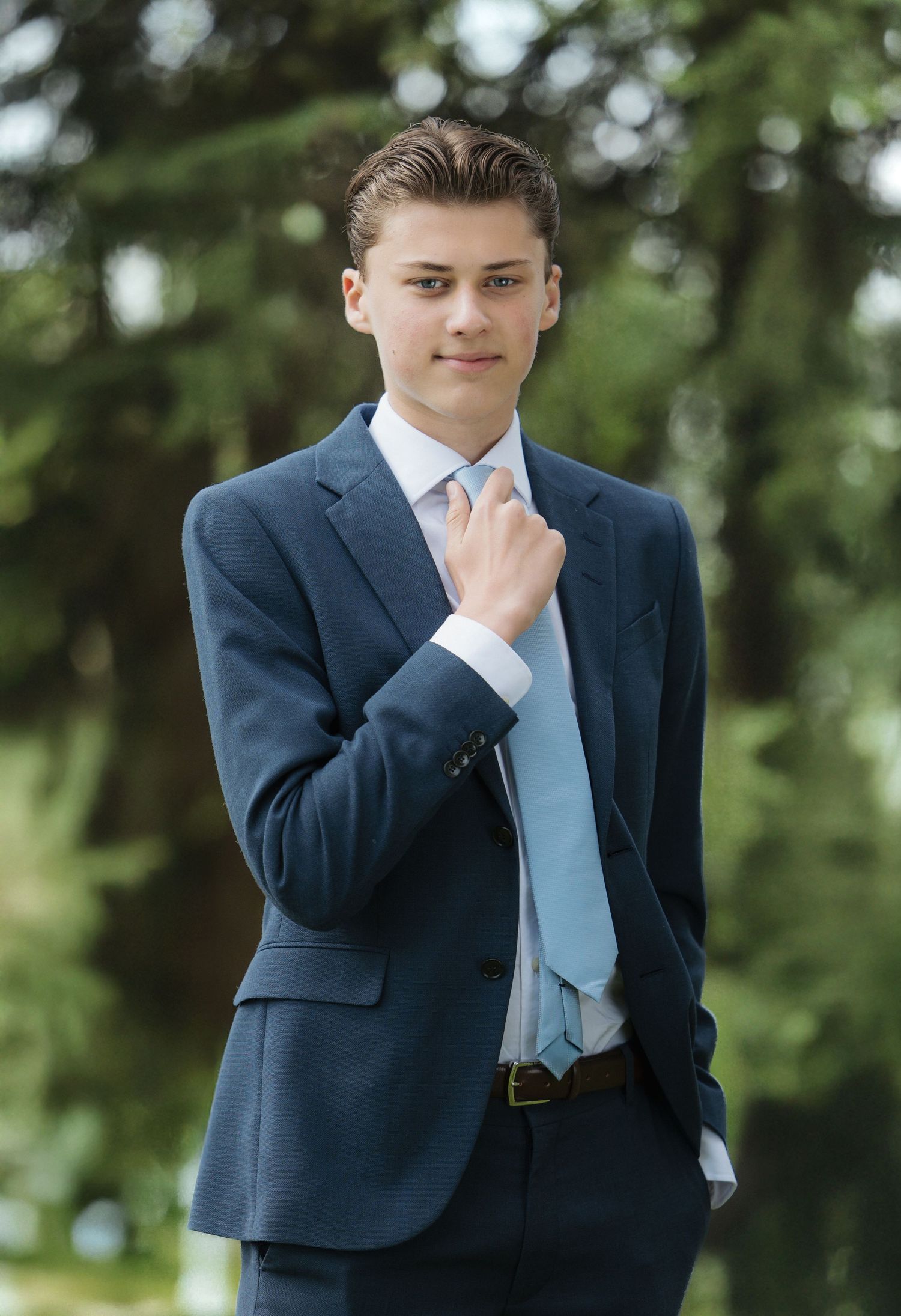 A person in a navy suit and light blue tie poses thoughtfully against a blurred green outdoor background.