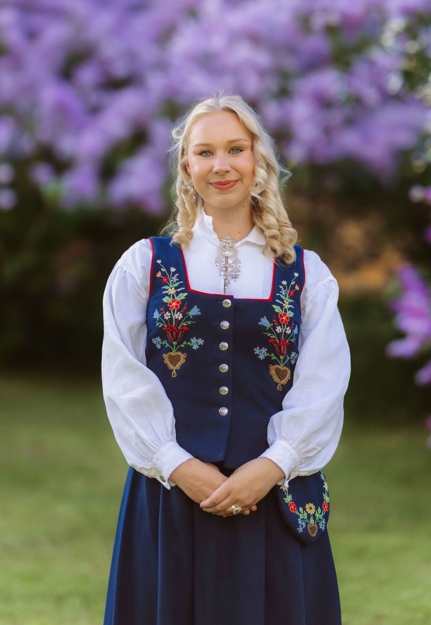 Young blonde woman in traditional Norwegian bunad folk costume with floral embroidery, posing outdoors near purple flowers.