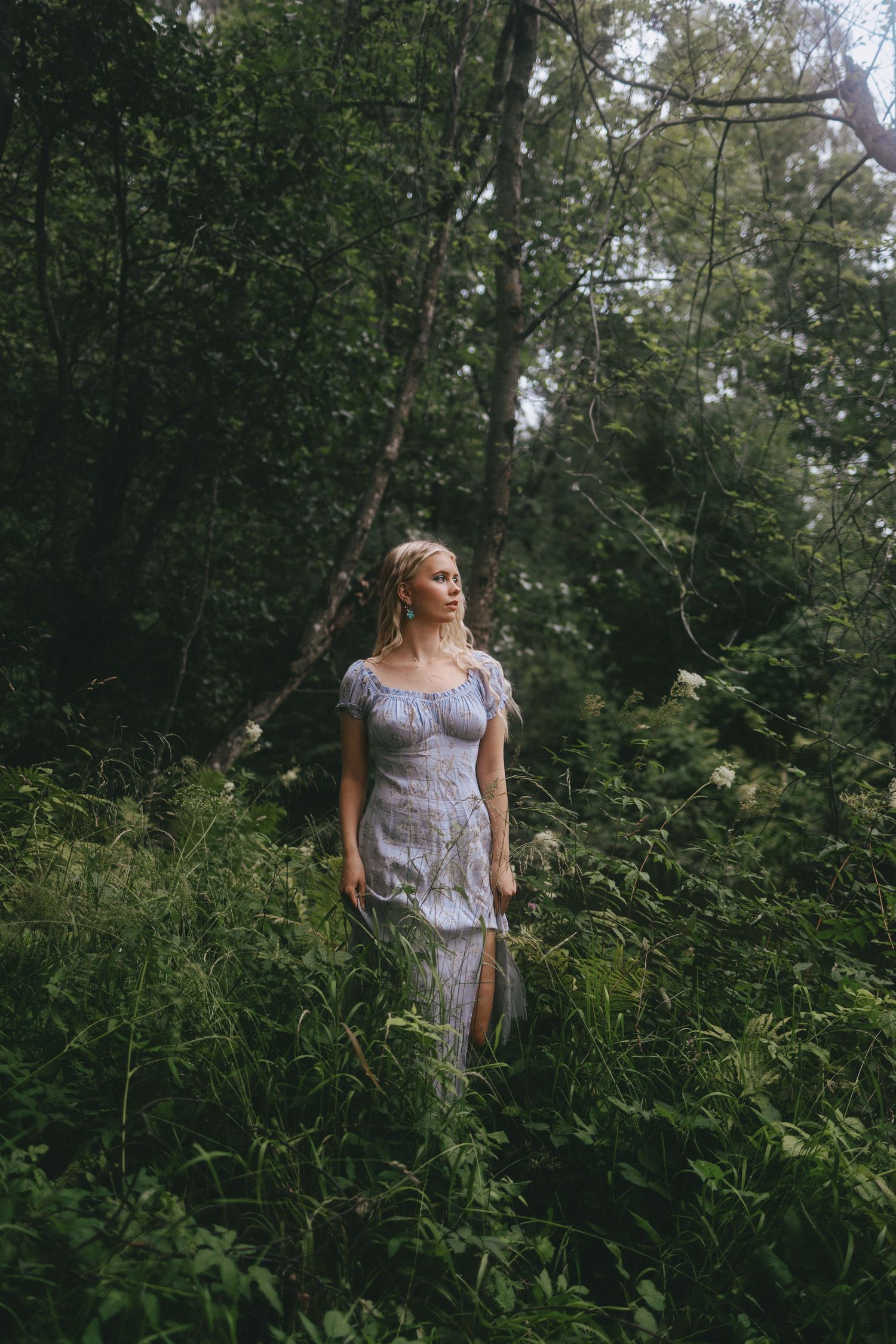 A person in a light purple dress stands among tall grass and trees in a dreamy forest setting.
