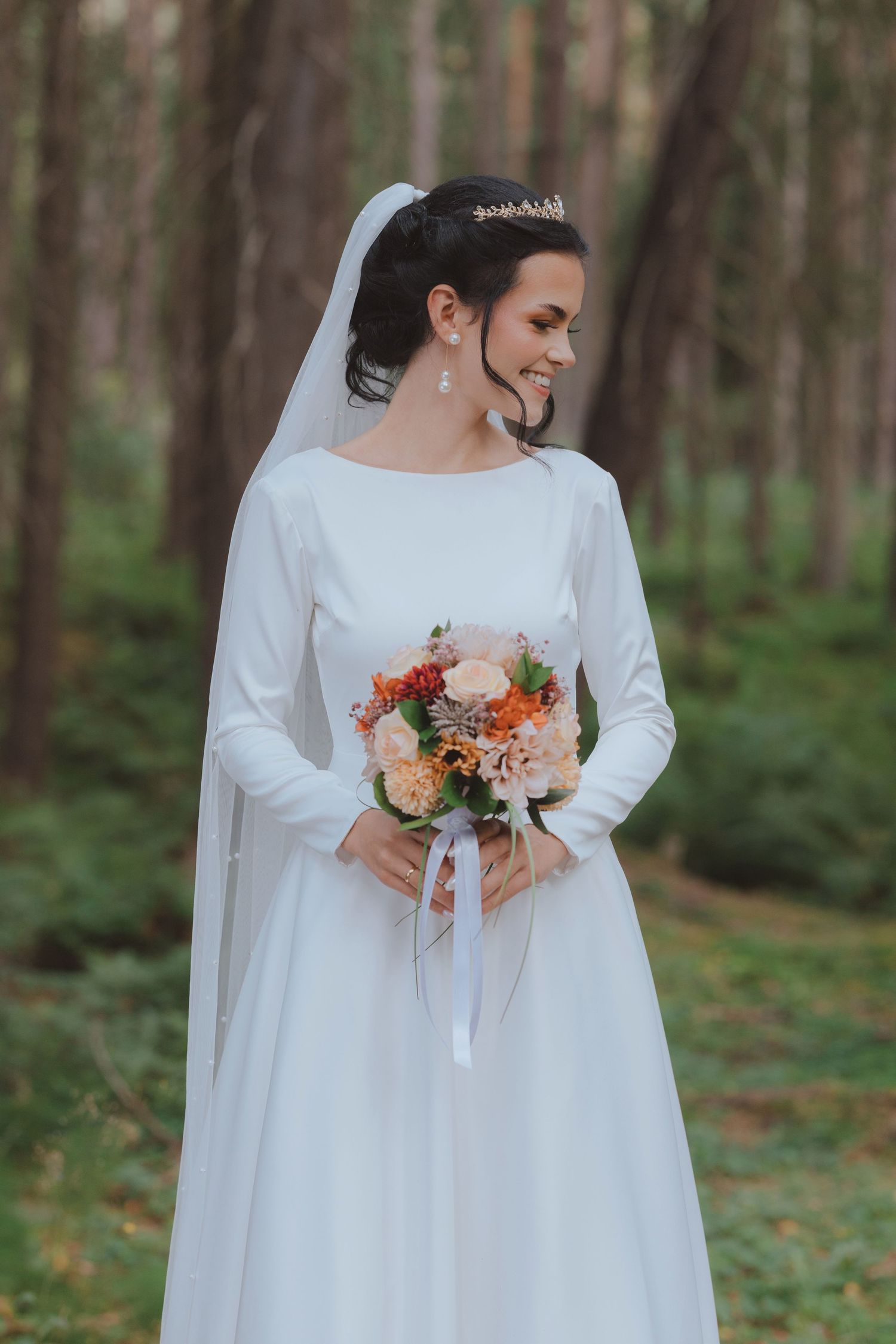 Bride in long-sleeved white wedding dress holds autumn bouquet in forest setting with orange and peach flowers.