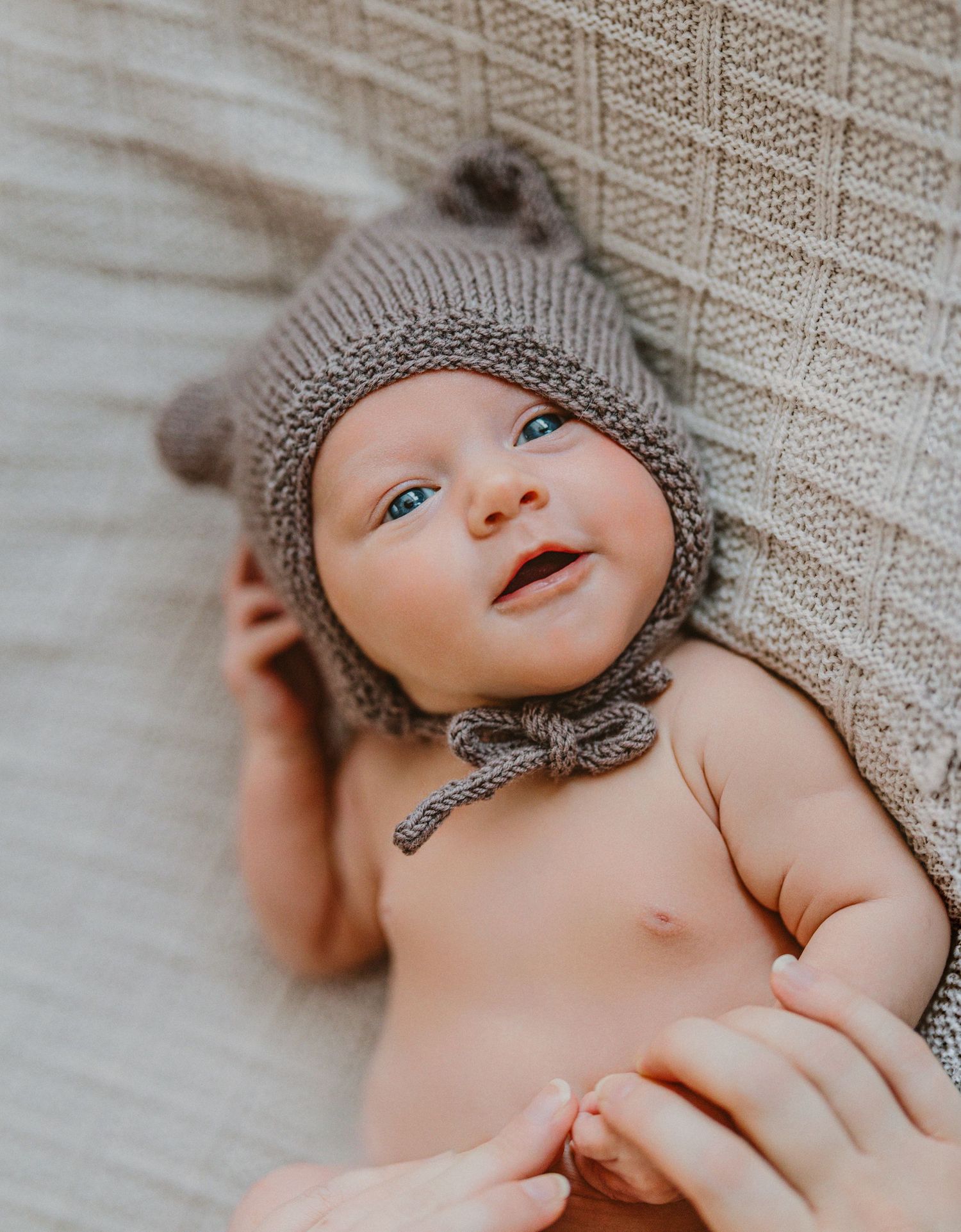 Baby wearing a gray knit bonnet lying on textured fabric with a sweet expression.