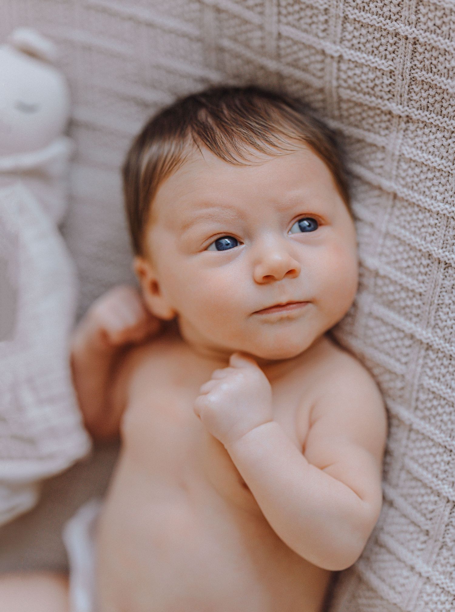 A baby lies on soft beige bedding while looking up with bright blue eyes and a curious expression.