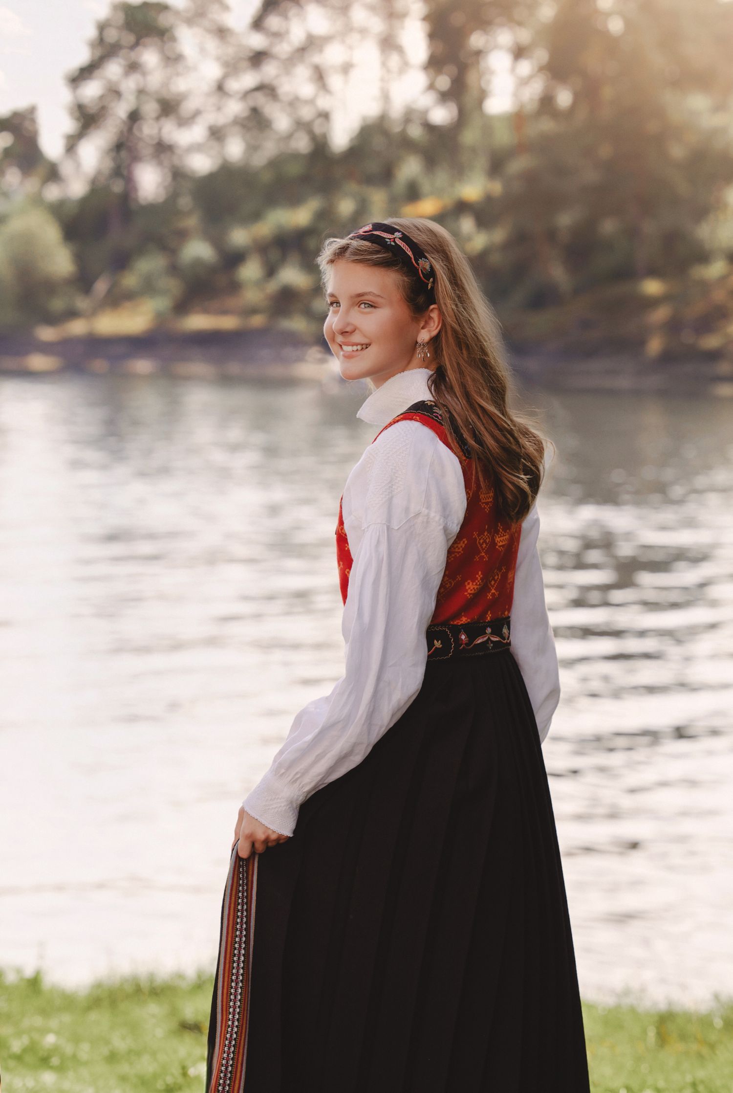Person wearing traditional Norwegian folk costume stands by tranquil lake at sunset in scenic outdoor setting.