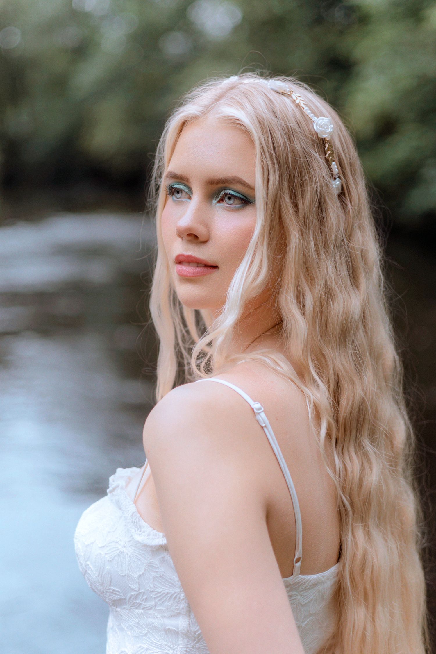 A woman in a white dress with blonde wavy hair poses outdoors against a blurred natural background.