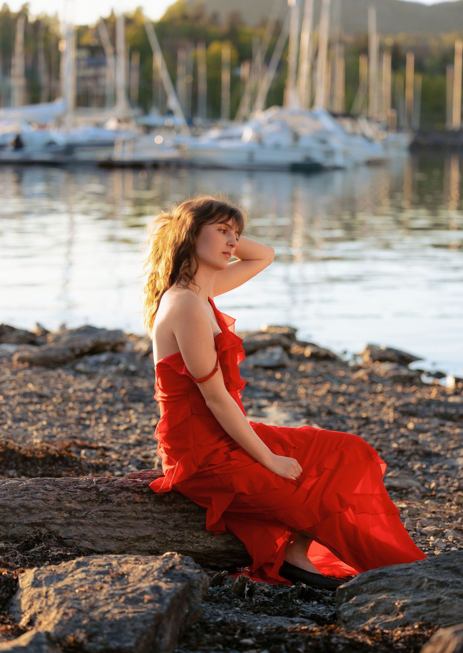 Person in flowing red dress poses gracefully by waterfront at sunset.