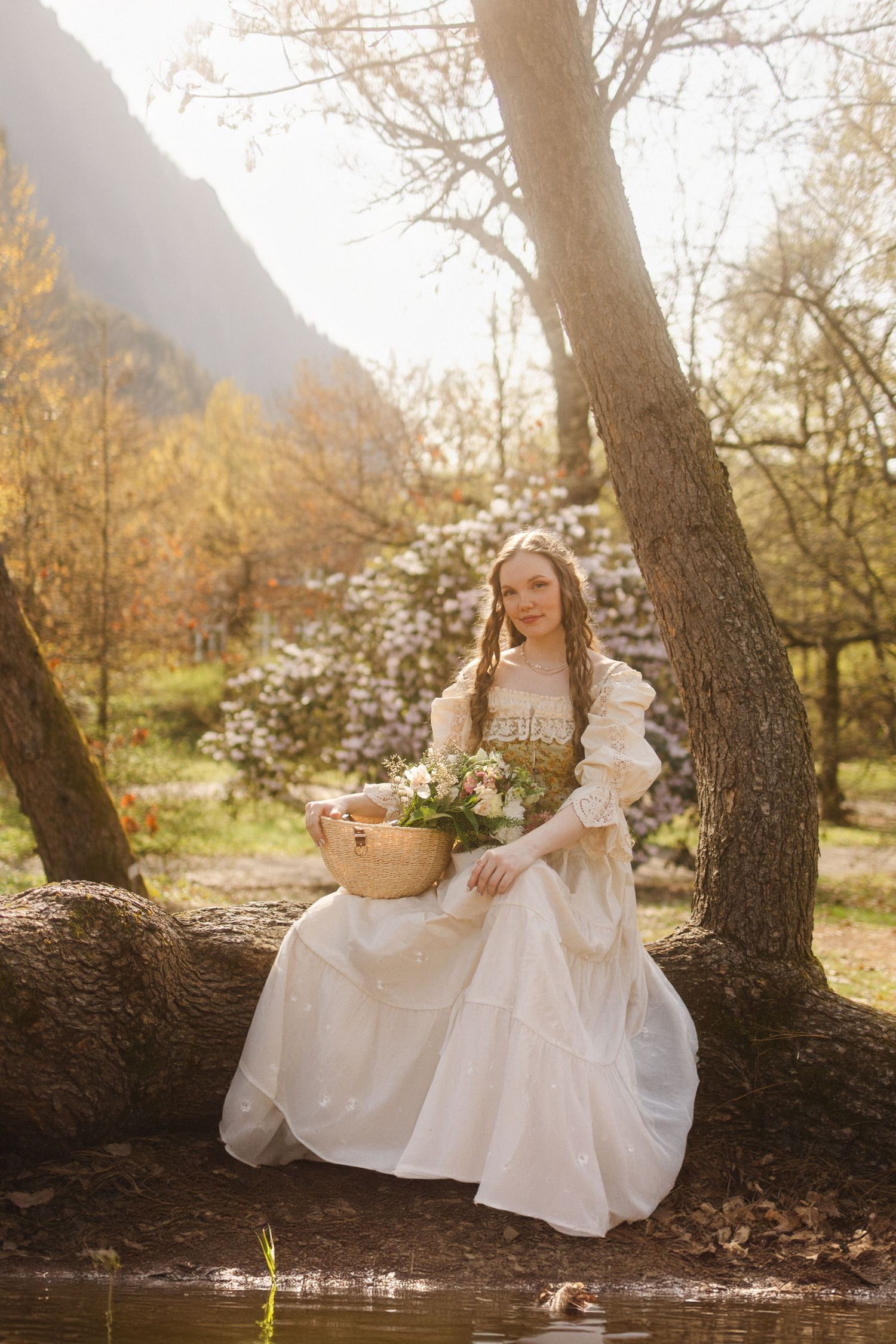 A dreamy outdoor portrait session features someone in an elegant white dress sitting beneath large trees in soft natural light.