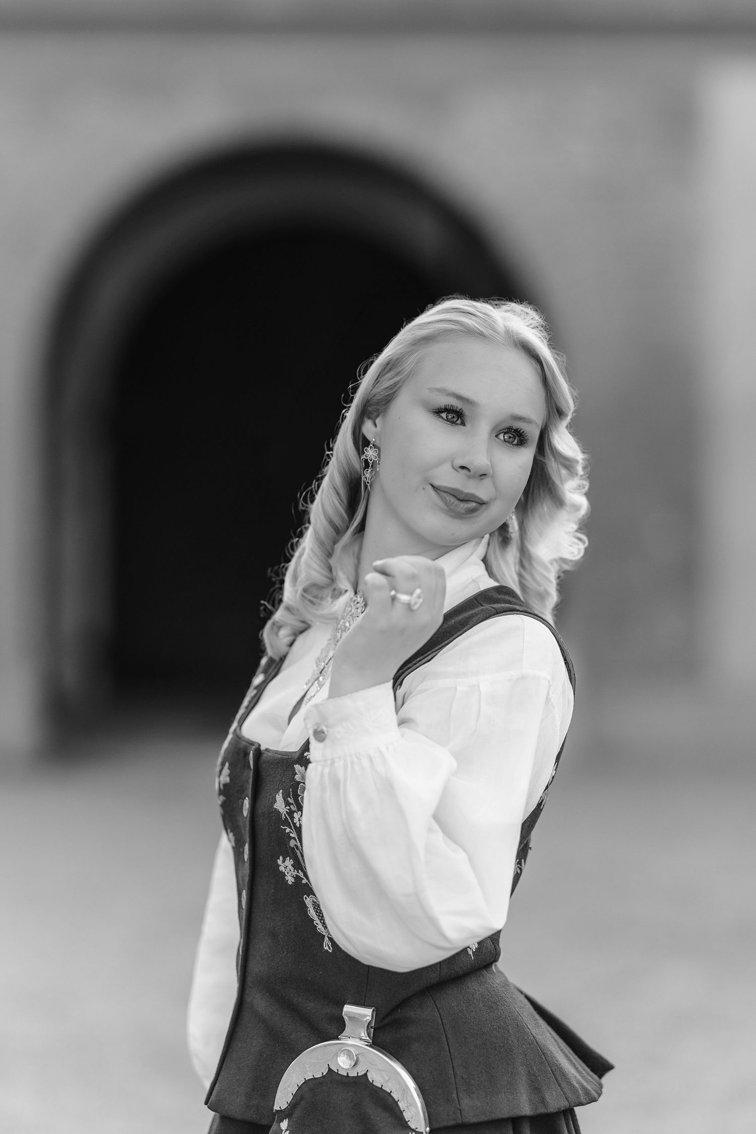 Black and white portrait of a blonde woman in traditional folk costume standing before an arched stone doorway.