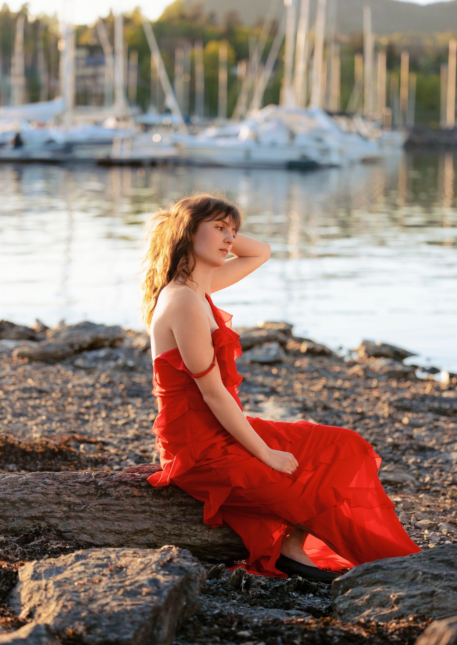 A woman in a flowing red dress sits on rocks by a marina at sunset.