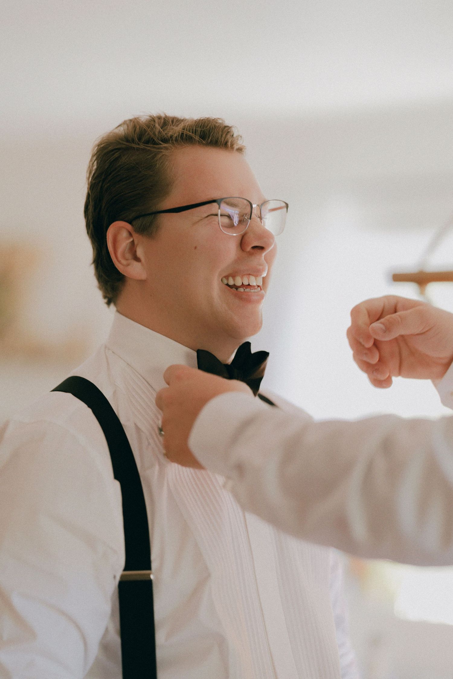 A person wearing a white dress shirt and black suspenders smiles while getting ready.
