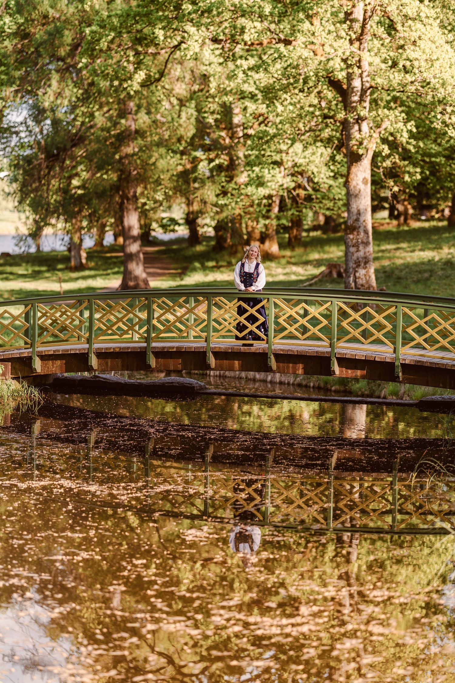 Woman in traditional dress standing on a wooden bridge over a calm pond in a lush green park.