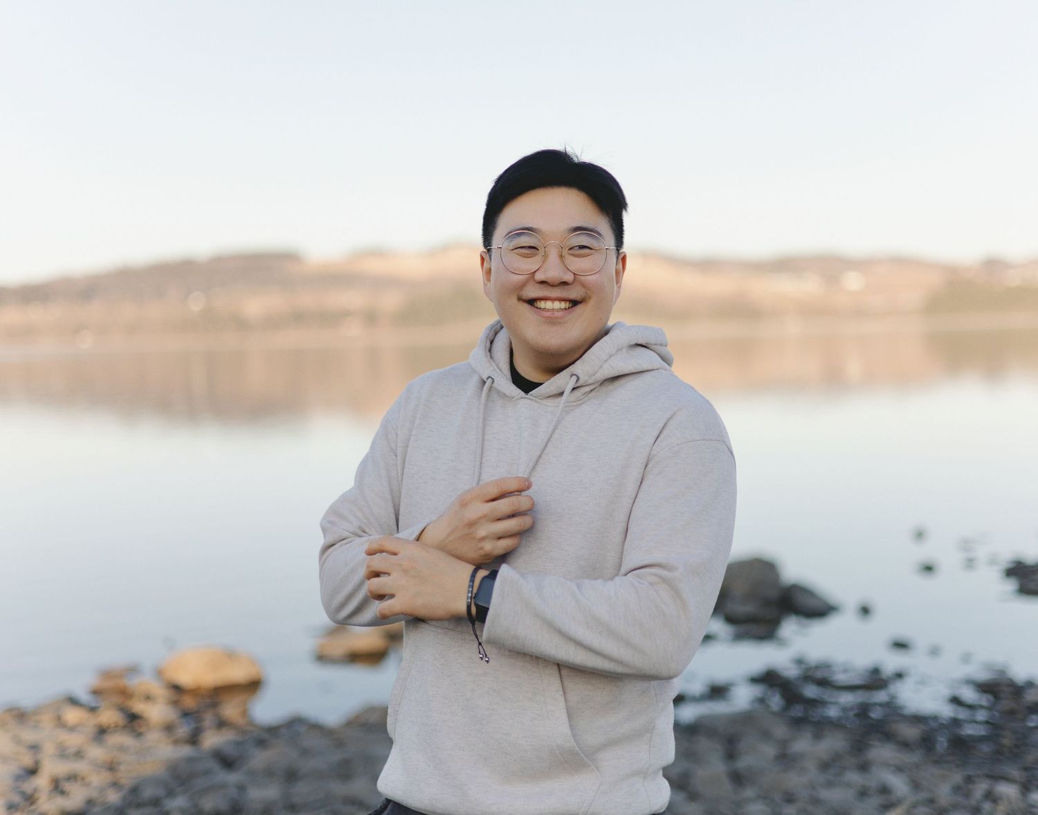 Someone wearing a light gray hoodie smiles while standing near a calm lake with rocky shores at sunset.